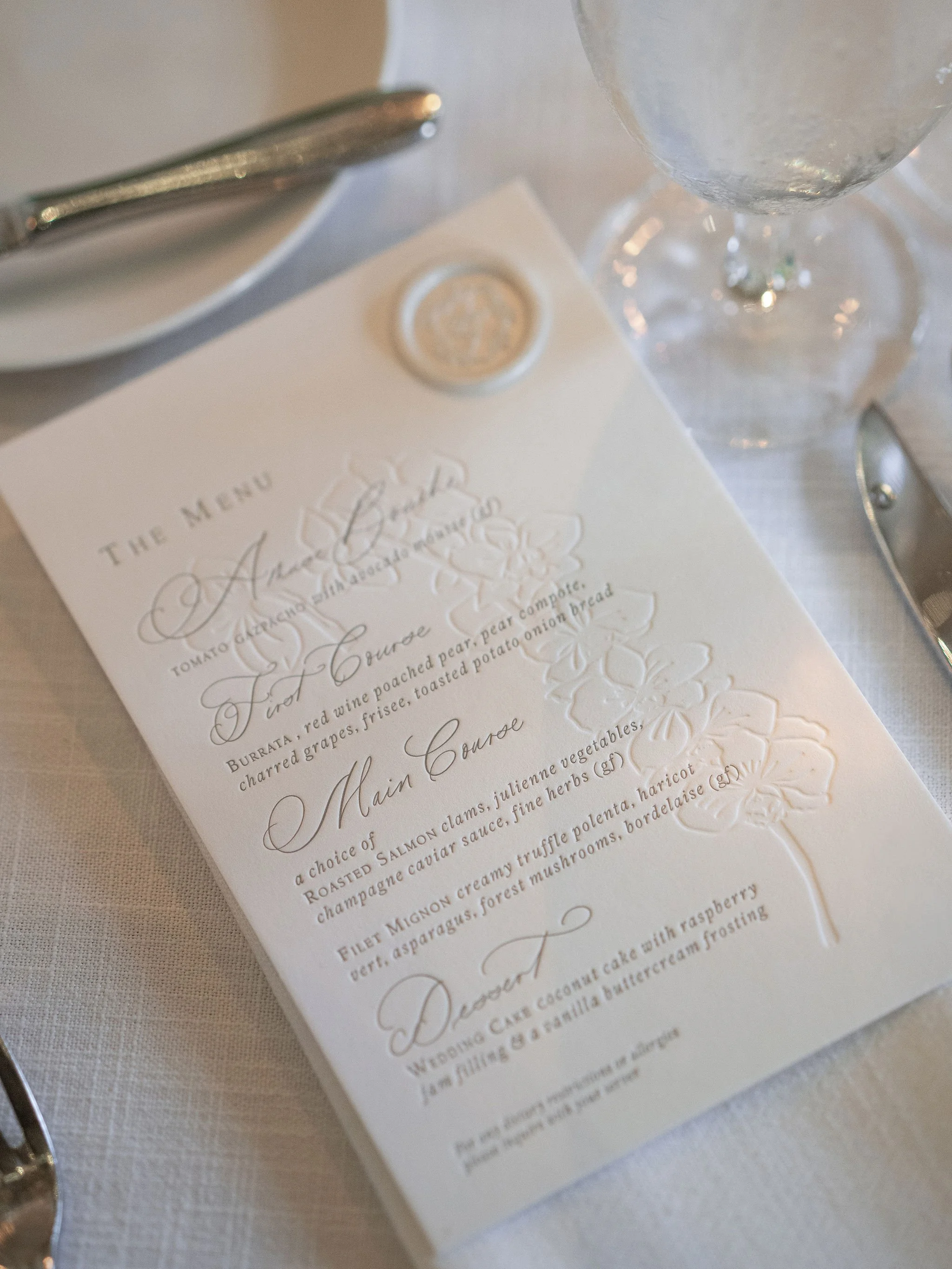 Elegant wedding menu with embossed floral design, featuring appetizers, main courses, and a dessert, placed on a white tablecloth next to glassware and silverware.
