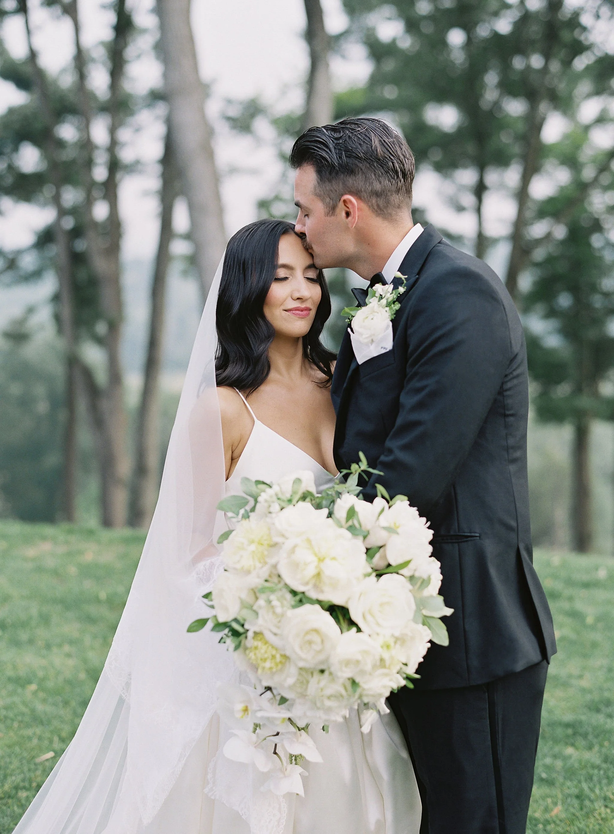 A bride and groom sharing a tender moment outdoors on their wedding day. The groom kisses the bride on her forehead while she holds a large bouquet of white roses and greenery. The bride wears a white dress with thin straps and has long dark hair, wh