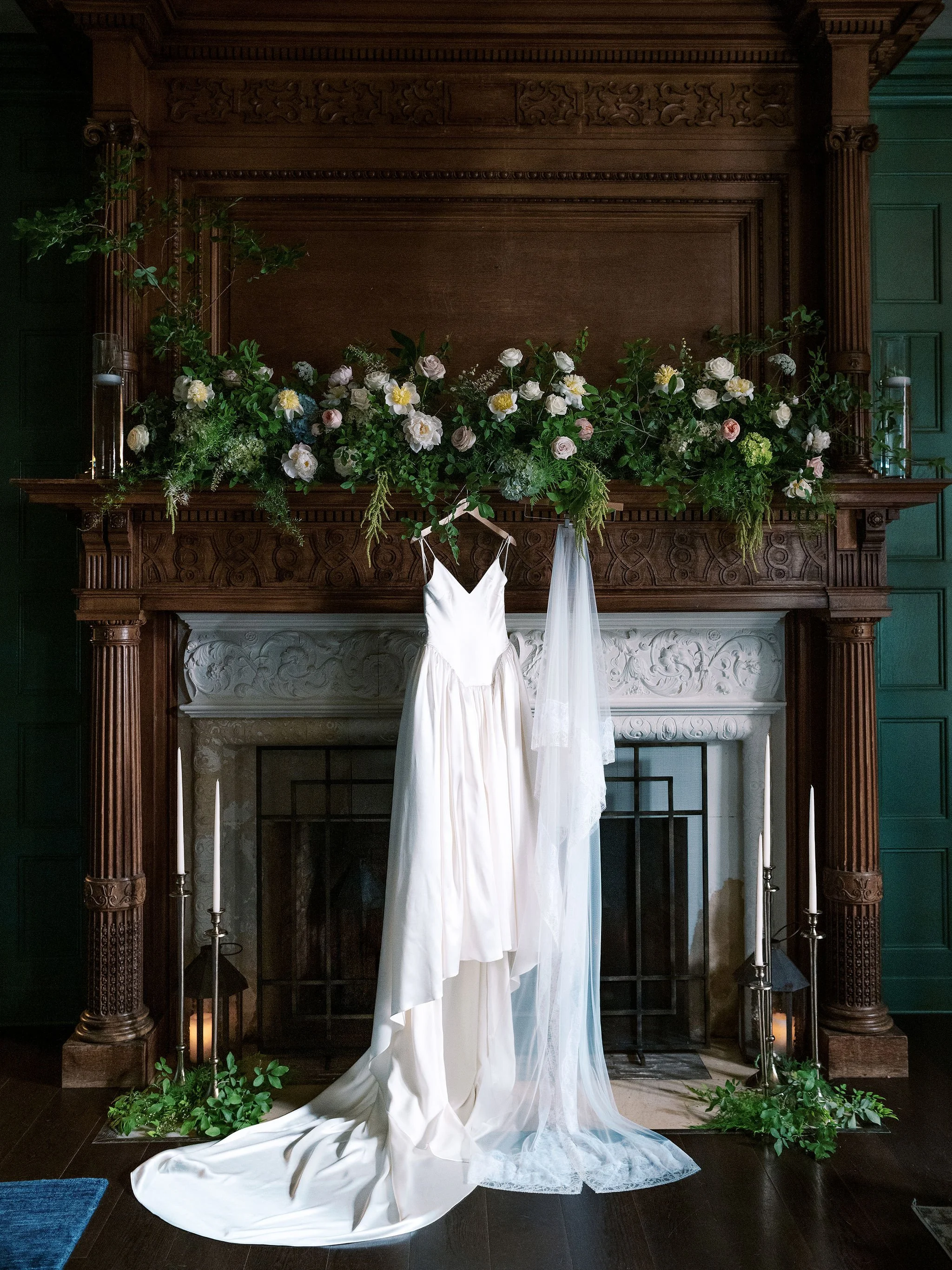 A wedding dress hanging in front of a decorated fireplace with a floral garland and candles.