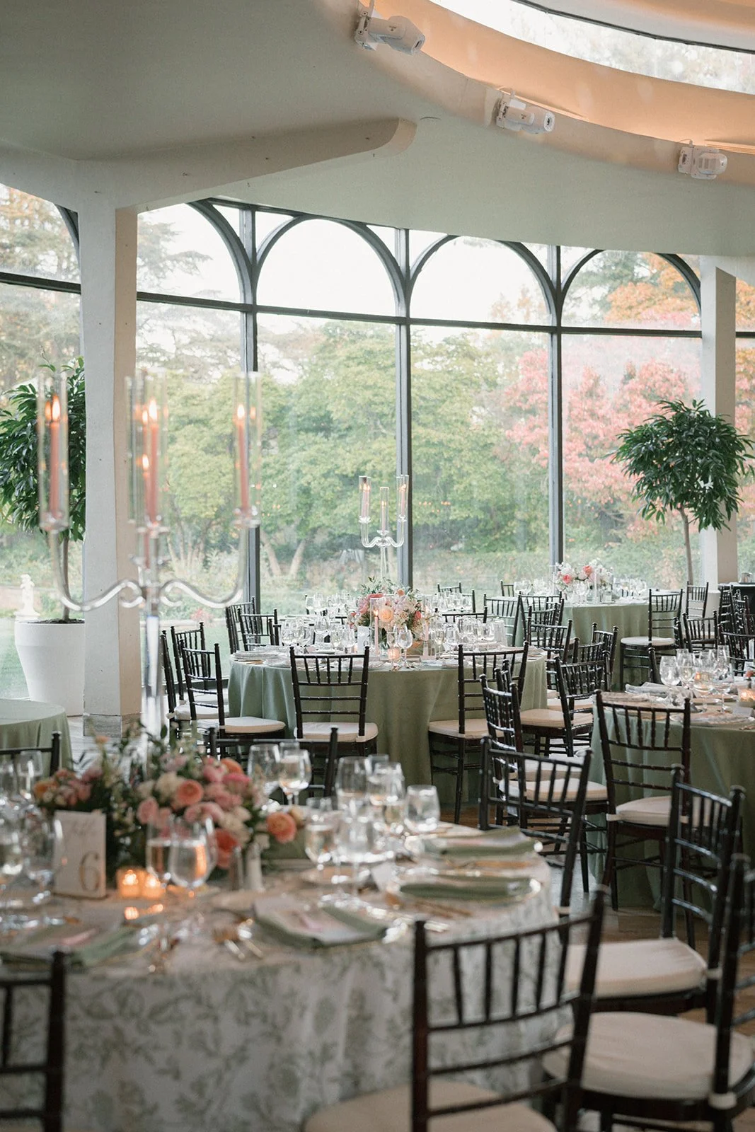 Elegant banquet hall decorated for a wedding or special event with round tables covered in green tablecloths, floral centerpieces, and multiple wine glasses, set in front of large windows showing a scenic outdoor view with trees and autumn foliage.