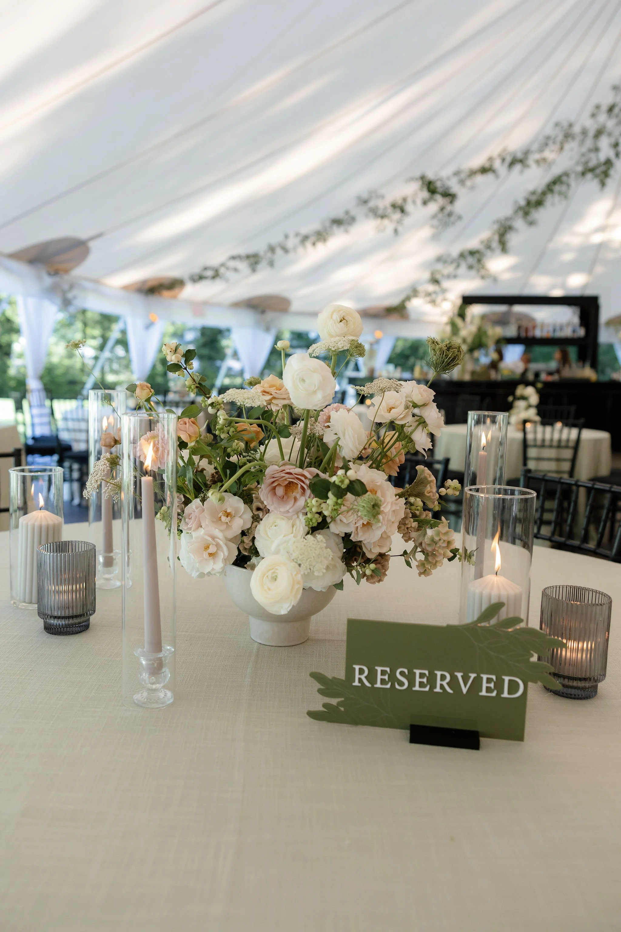 A floral centerpiece on a reserved wedding table includes white and blush pink flowers in a white vase, surrounded by candles in glass holders and a green reserved sign.