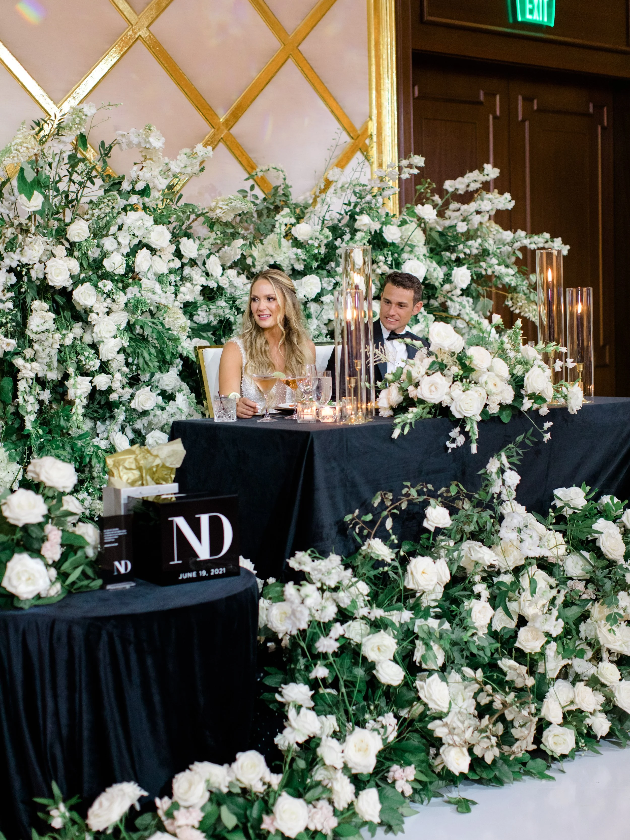 Bride and groom sitting at a wedding reception table, decorated with white flowers and tall candles, smiling and celebrating.
