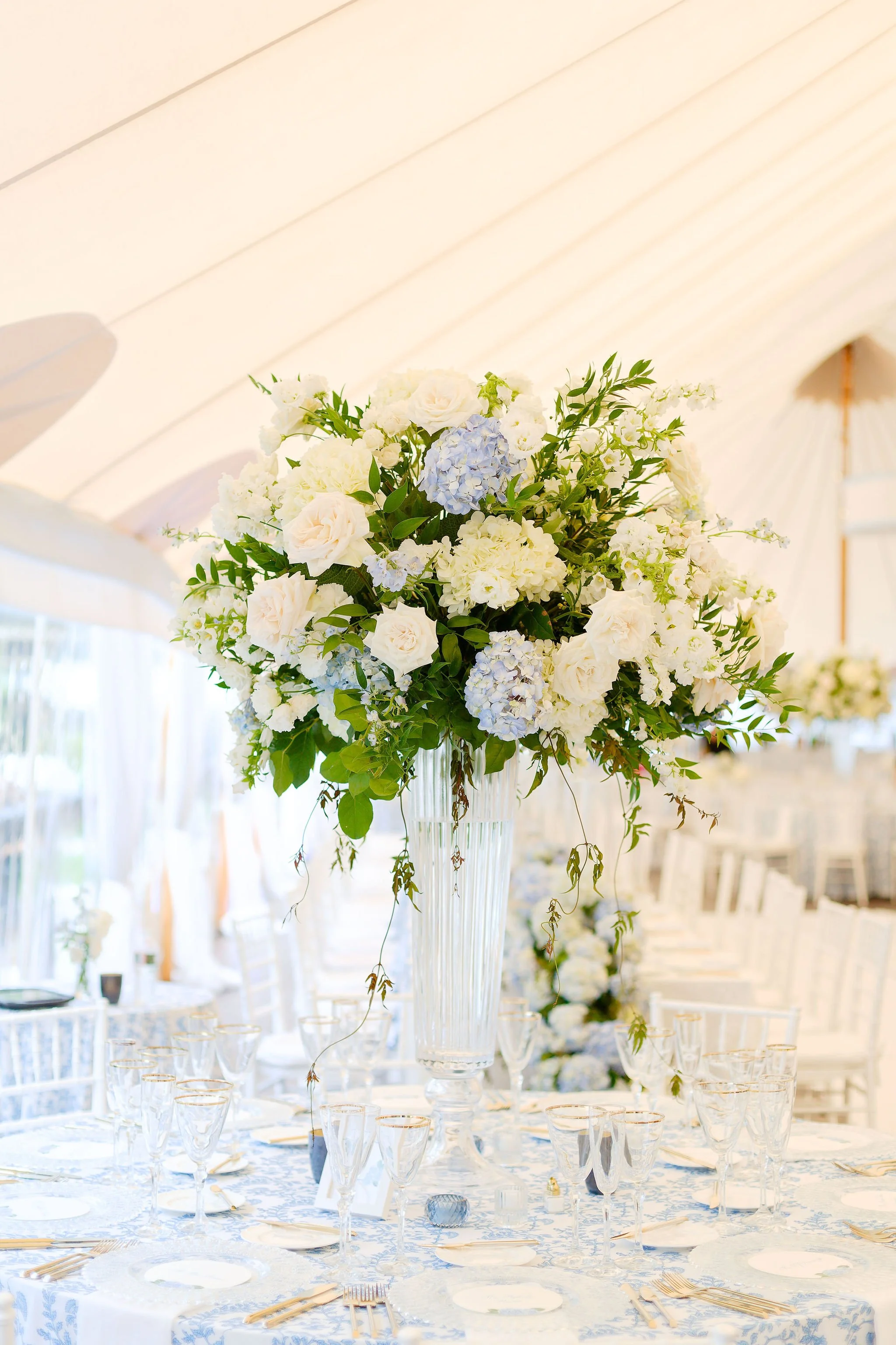 A tall glass vase with a large bouquet of white and light blue flowers on a wedding or event table with glassware and cutlery.