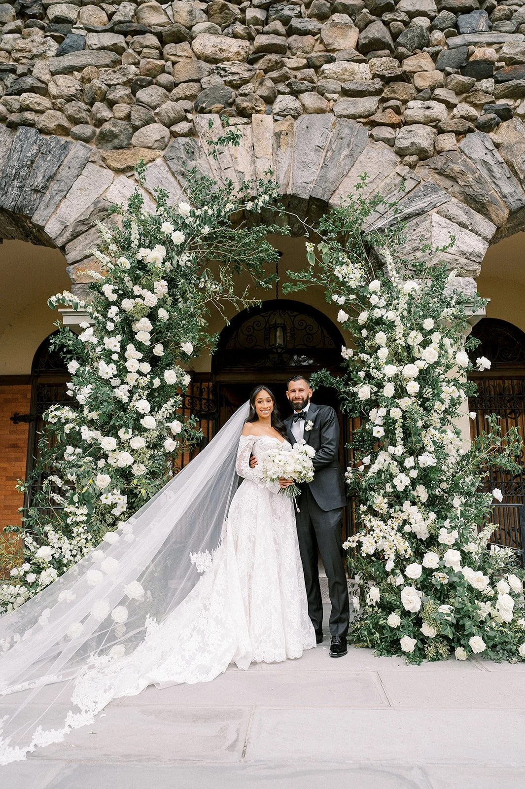 A bride and groom standing under a floral archway on their wedding day.