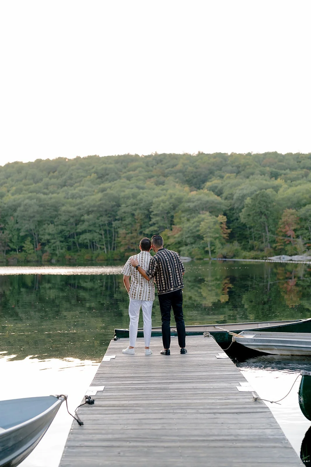 Two men standing on a wooden dock by a lake with a forested hill in the background, one has his arm around the other, both wearing patterned shirts.
