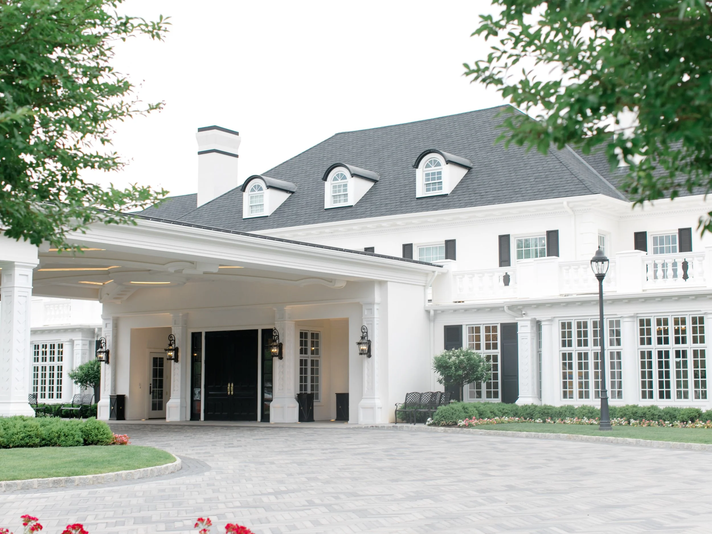 A large white mansion with black shutters, a gray shingled roof and a driveway leading to a black front door, surrounded by landscaping and trees.