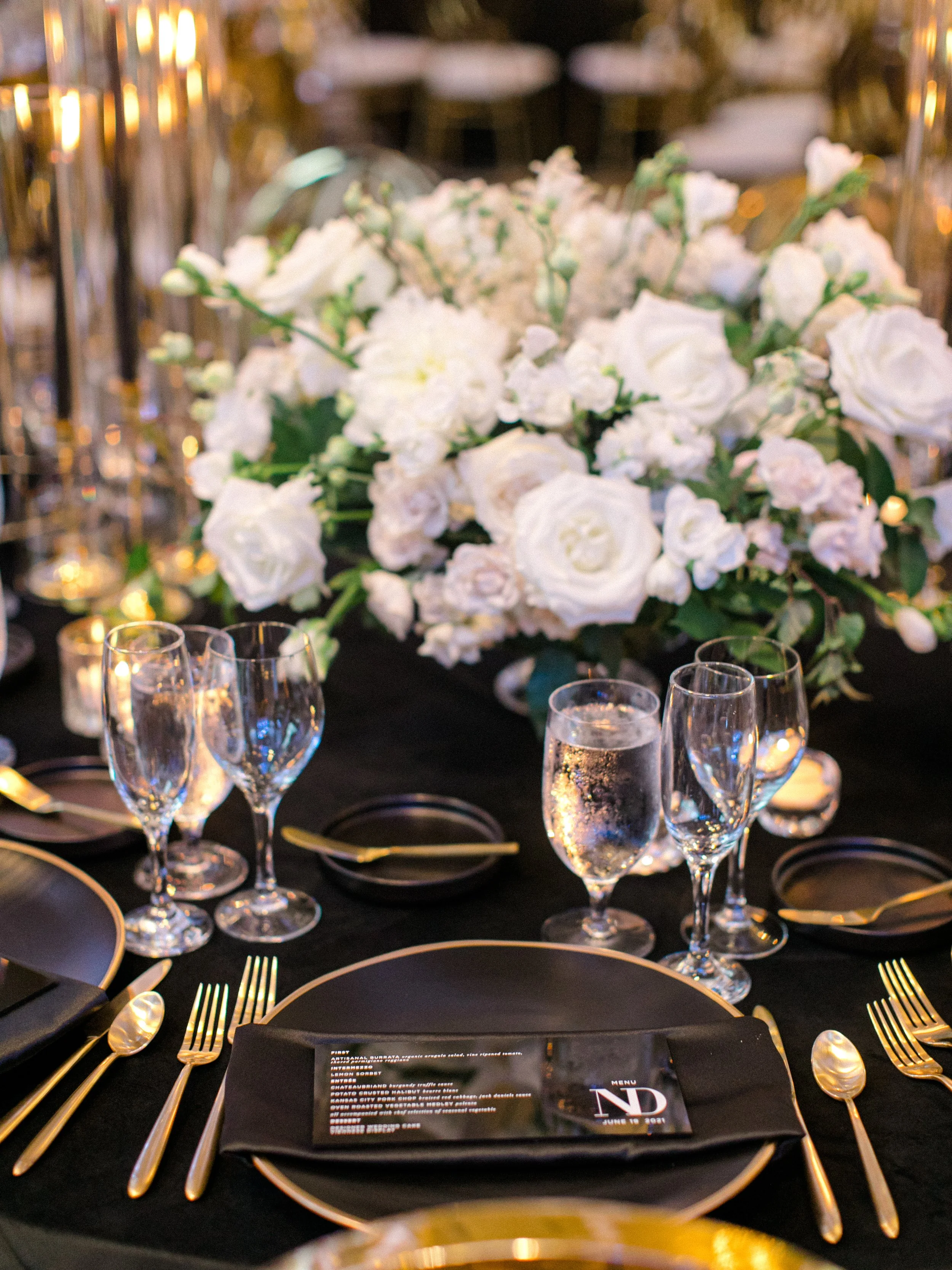 Elegant table setting with black tablecloth, gold utensils, black plates with gold rims, champagne flutes, a large floral centerpiece of white roses and flowers, and a menu card on the plate.