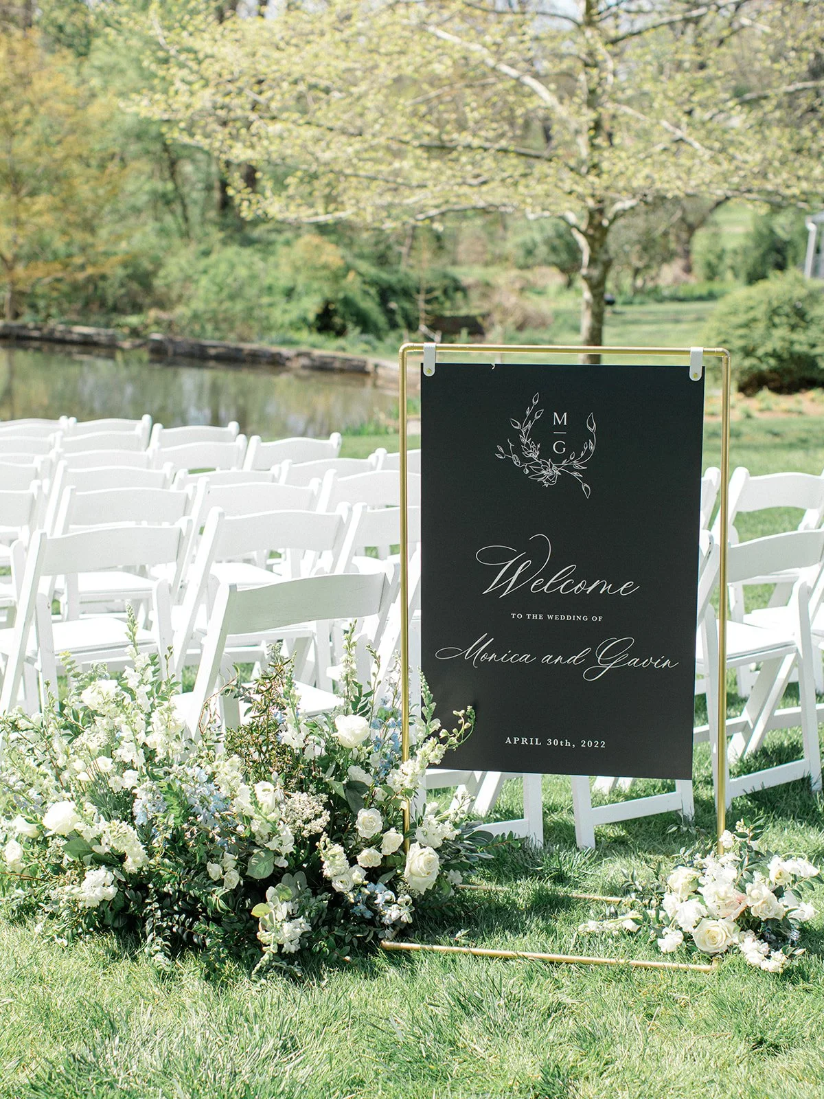 Outdoor wedding setup with white chairs, floral arrangement, and a black welcoming sign for Monica and Gavin's wedding on April 30th, 2022, by a pond with trees in the background.