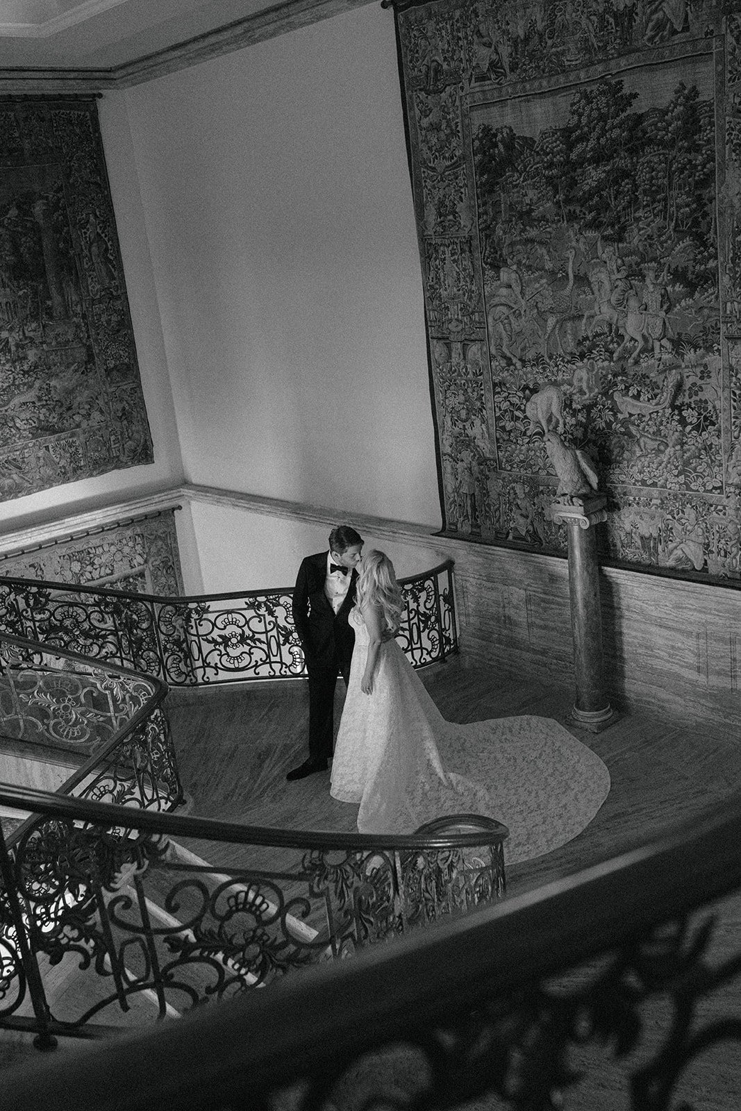 A black and white photograph of a bride and groom on a staircase in an elegant, vintage interior. The groom is wearing a tuxedo and bow tie, and the bride is in a wedding gown. They are looking at each other closely, surrounded by ornate decorative r
