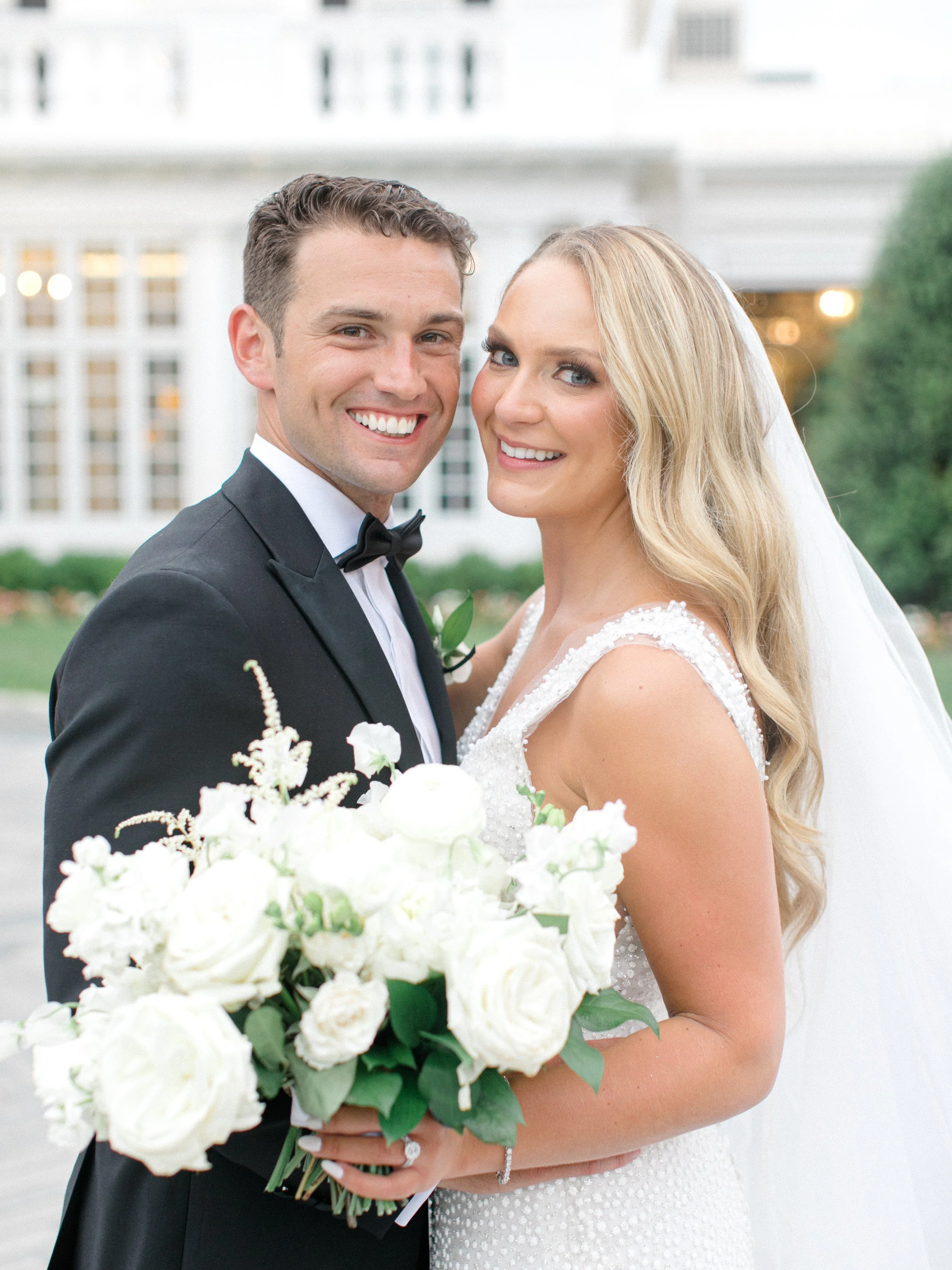 A happy bride and groom on their wedding day, standing outdoors, with the bride holding a bouquet of white flowers and wearing a white dress, and the groom in a black tuxedo with a bow tie, smiling at the camera.
