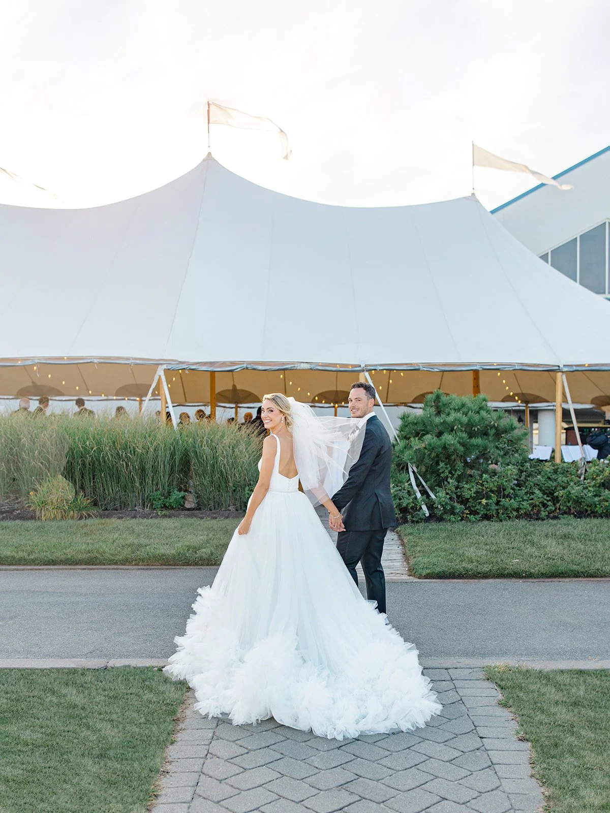 Bride and groom holding hands outside a wedding tent, bride in a white gown and veil, groom in a black suit, on a paved sidewalk with a grassy area and trees in the background.