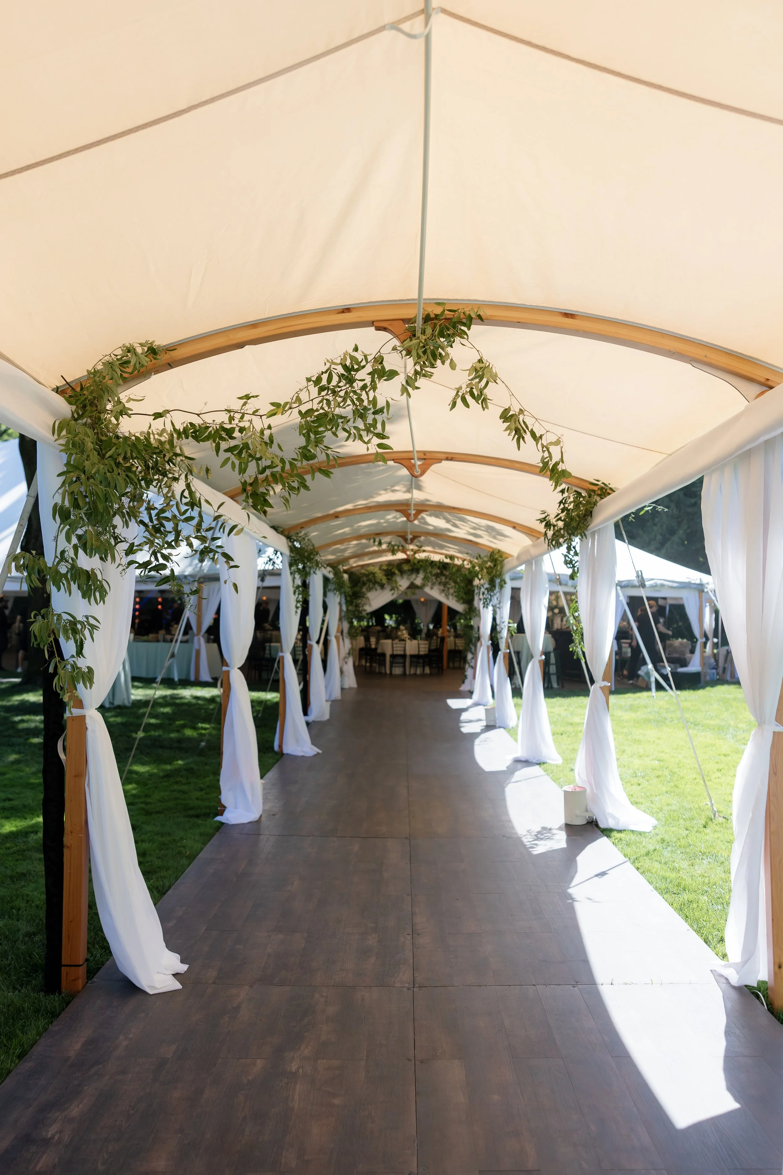 Decorated outdoor wedding venue with a canopy, white drapes, greenery, and a wooden walkway.