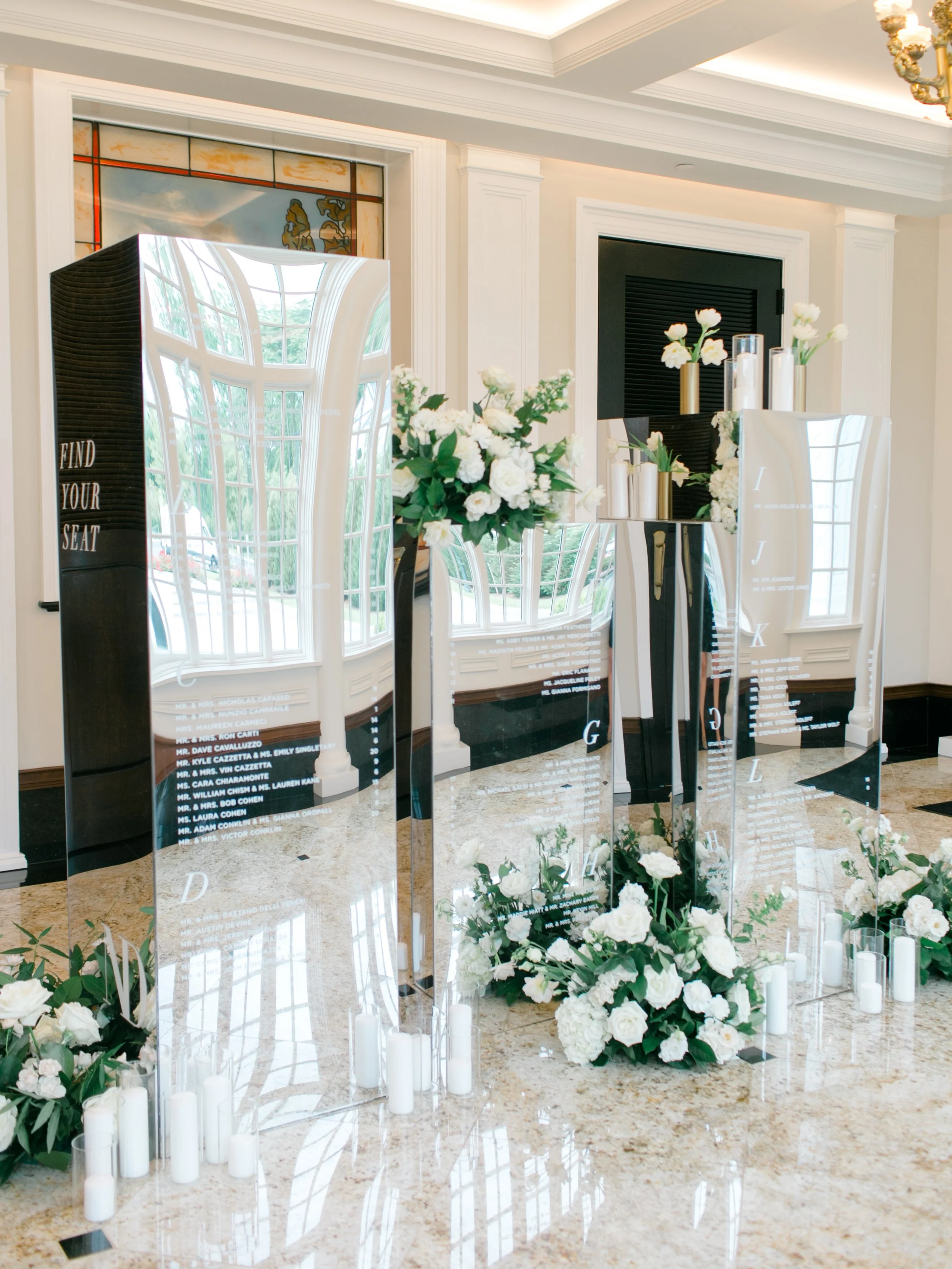Elegant wedding seating chart display with mirrored panels, floral arrangements, and white candles in a bright, stylish room.