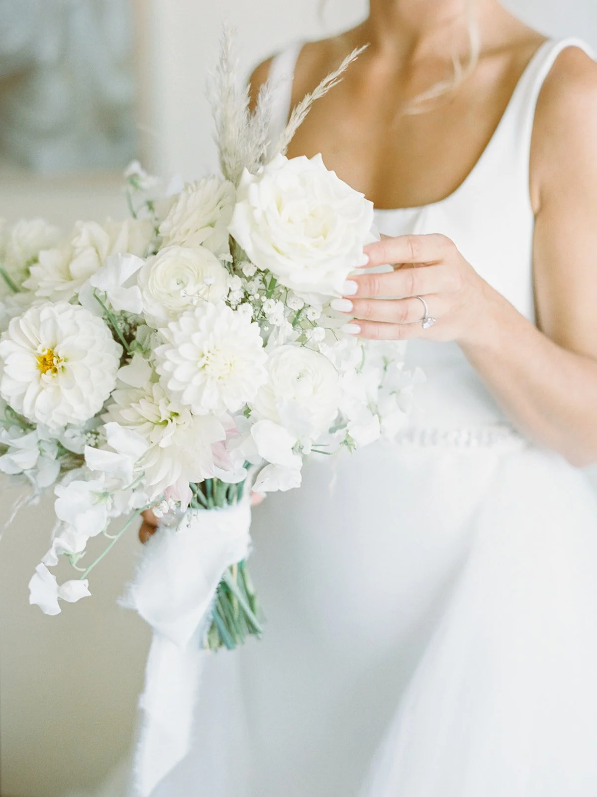 A woman in a white dress holding a bridal bouquet of white flowers, including roses and dahlias, with a ring on her finger.