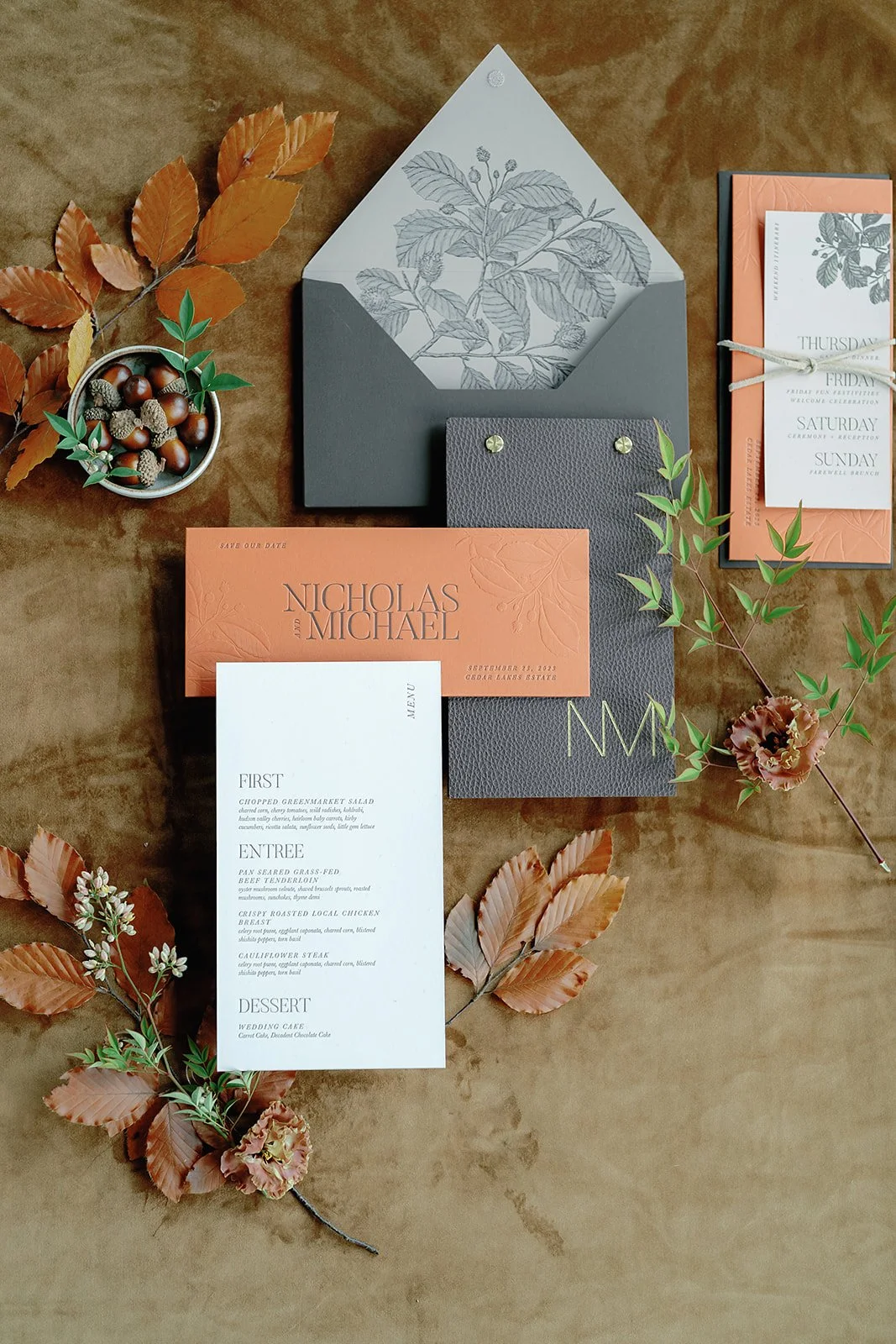 Wedding invitation suite with peach and gray color scheme, autumn leaves, acorns, berries, and a flower boutonniere on a wooden surface.