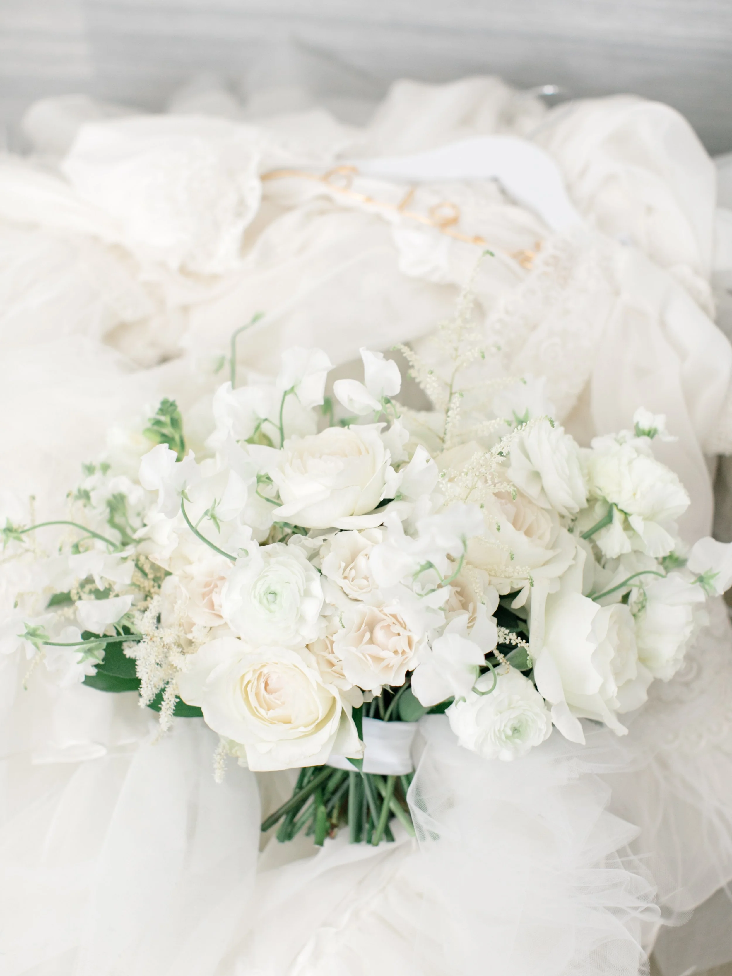 A white floral bouquet with roses and other white flowers, wrapped in white fabric, resting on a surface.