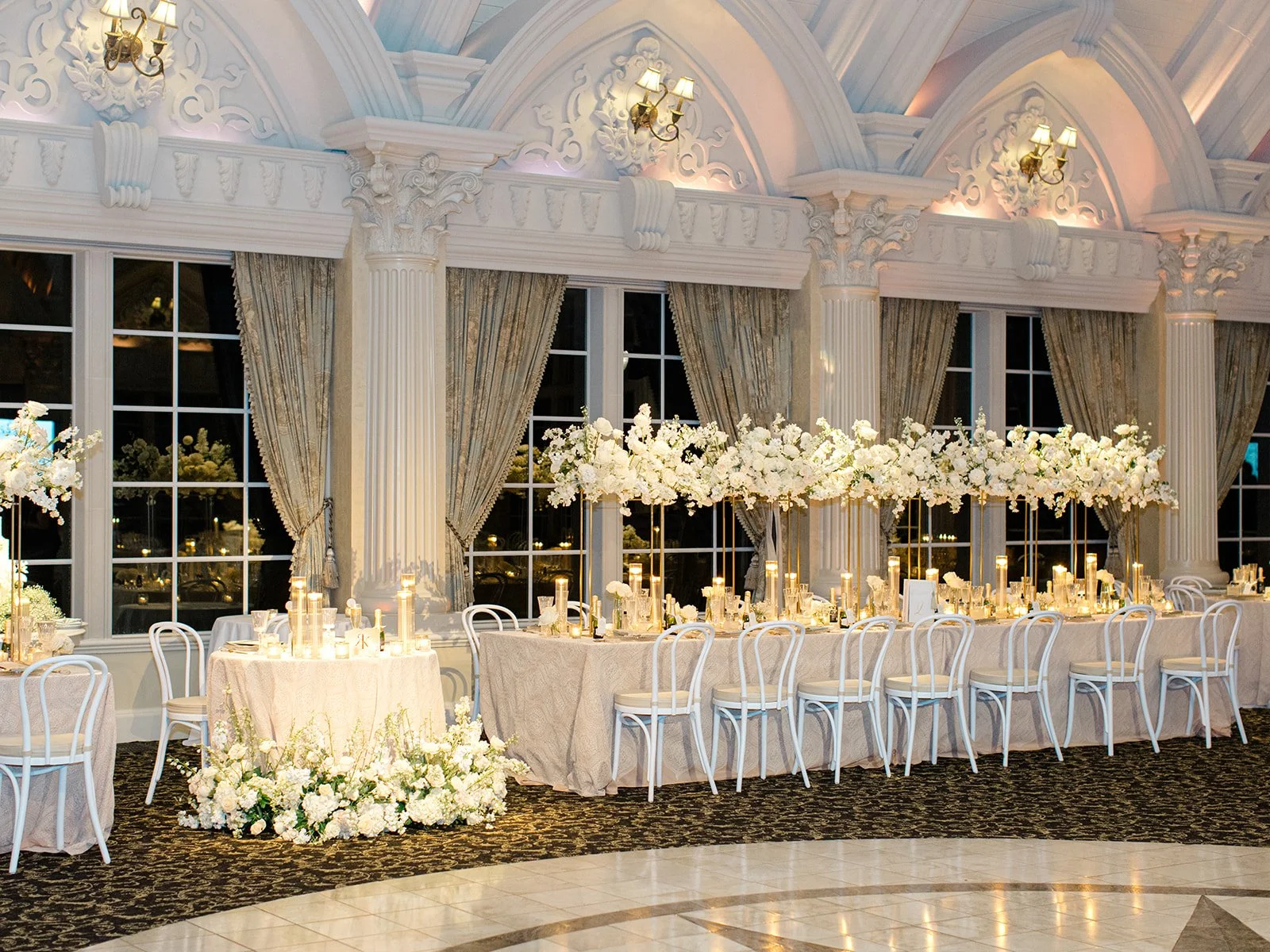 Elegant event space with long banquet table decorated with white flowers and candles, surrounded by white chairs, with large windows and ornate white architecture, including columns and archways.
