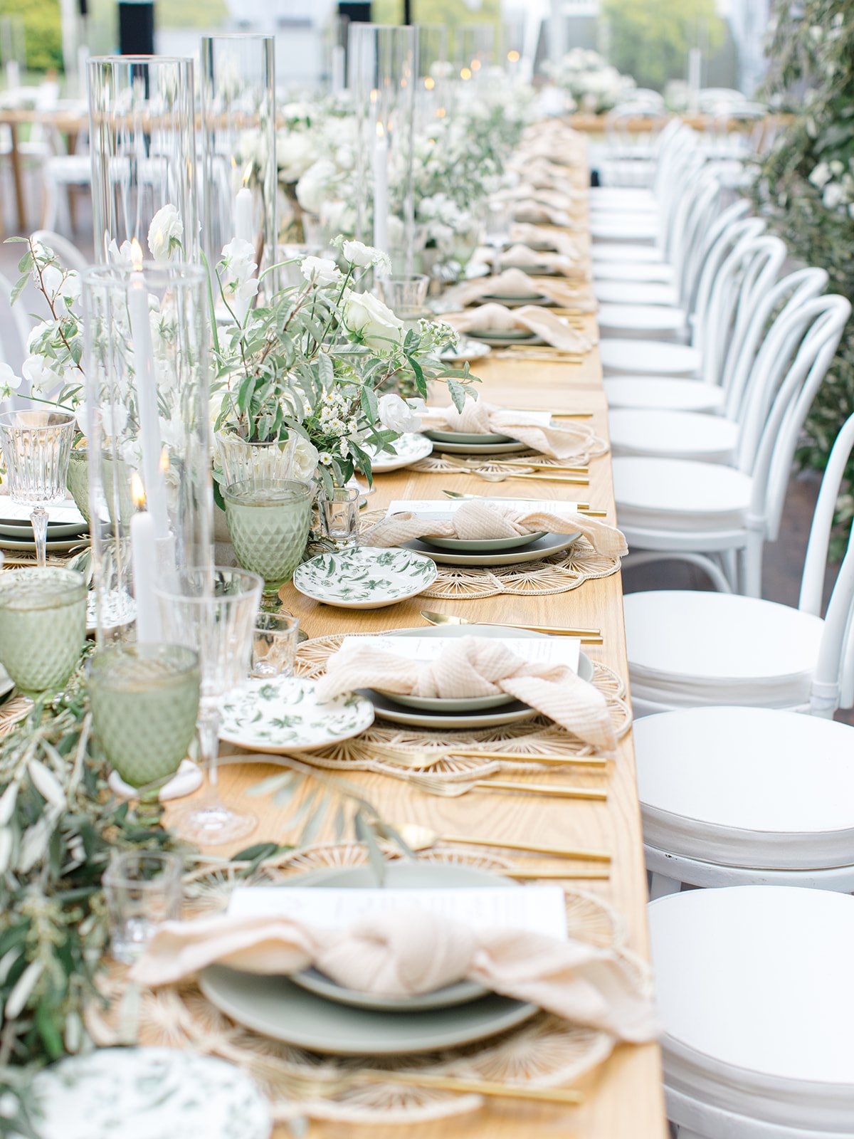 A long outdoor table set for a celebration with white chairs, green and white floral arrangements, candles, and elegant tableware.