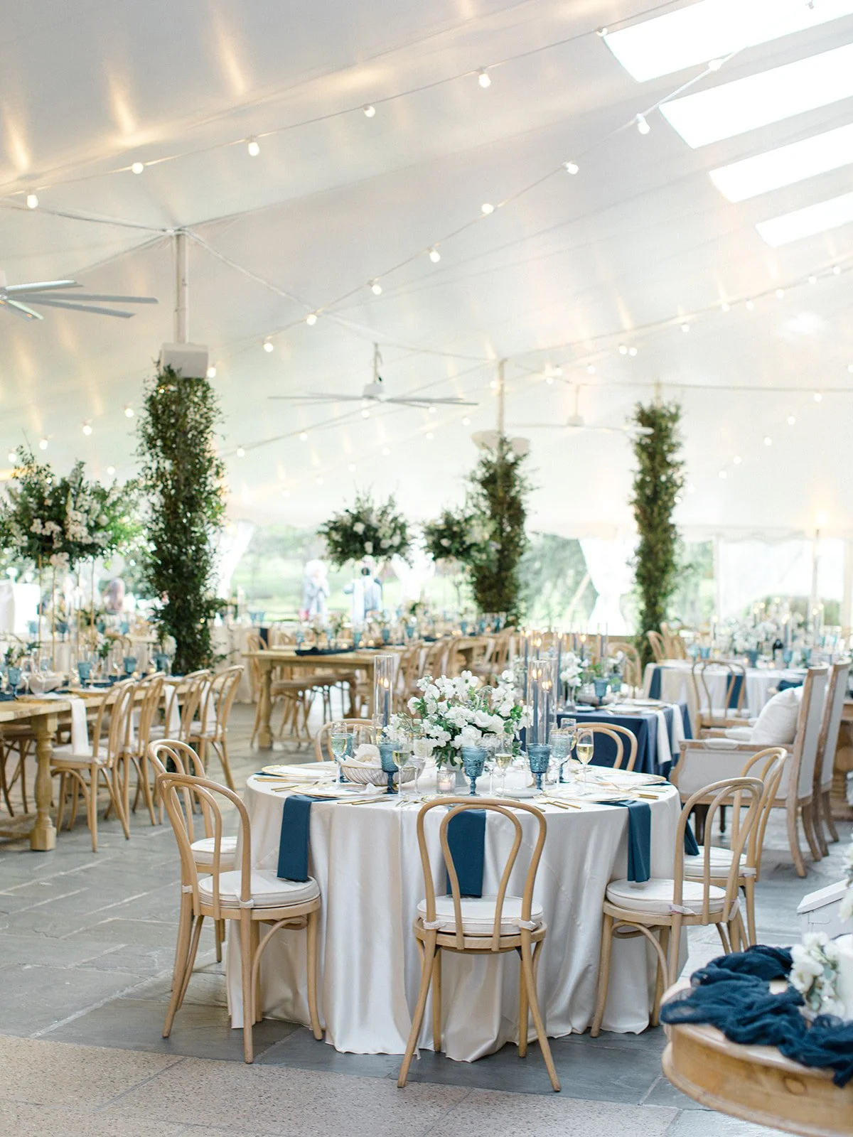 Elegant event tent with round tables decorated with white and blue table settings, floral centerpieces, and string lights hanging from the ceiling.