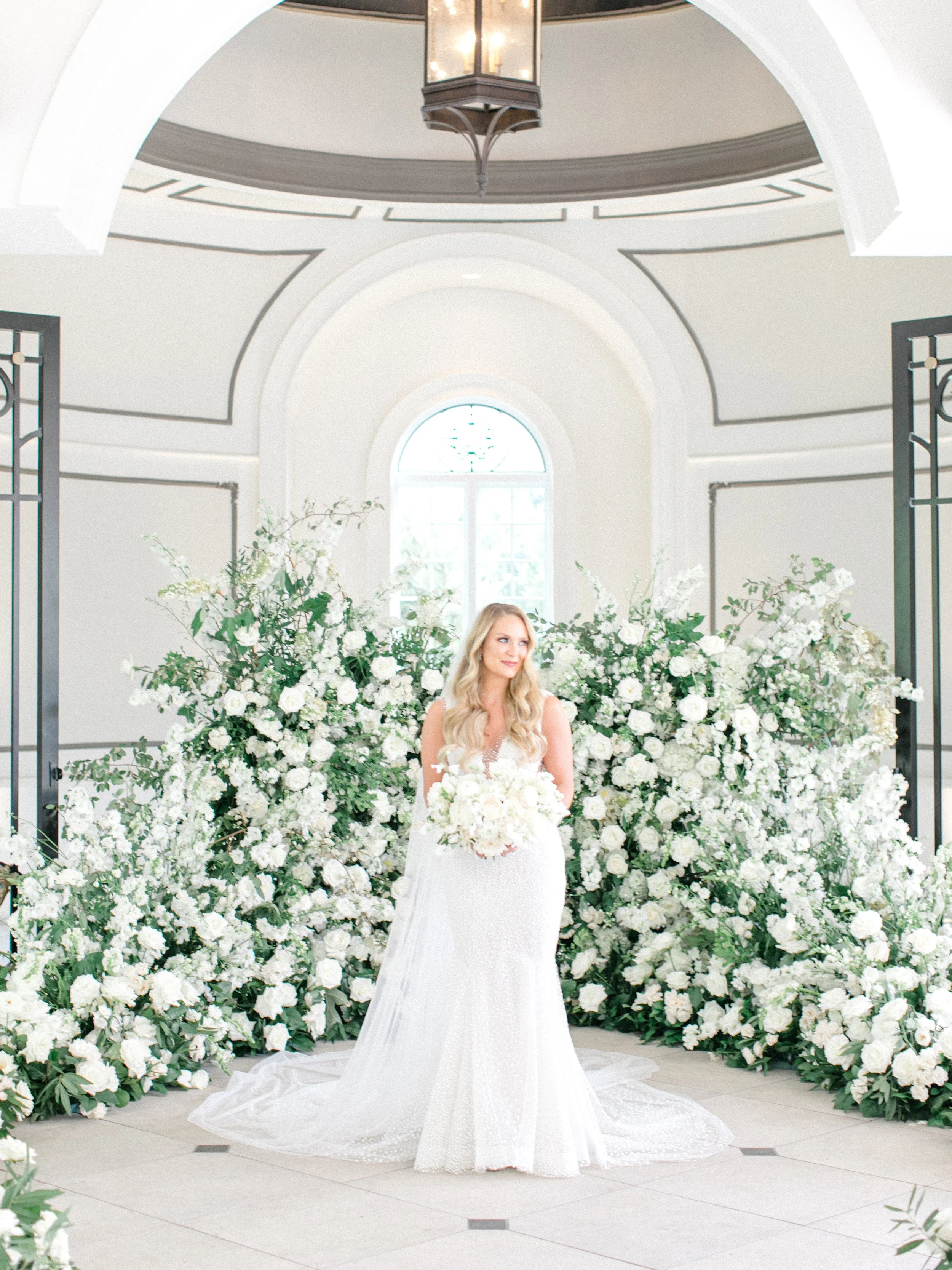 Bride in a white wedding dress holding a bouquet, standing in front of a large arrangement of white flowers and greenery, inside a building with arched windows and a high ceiling.