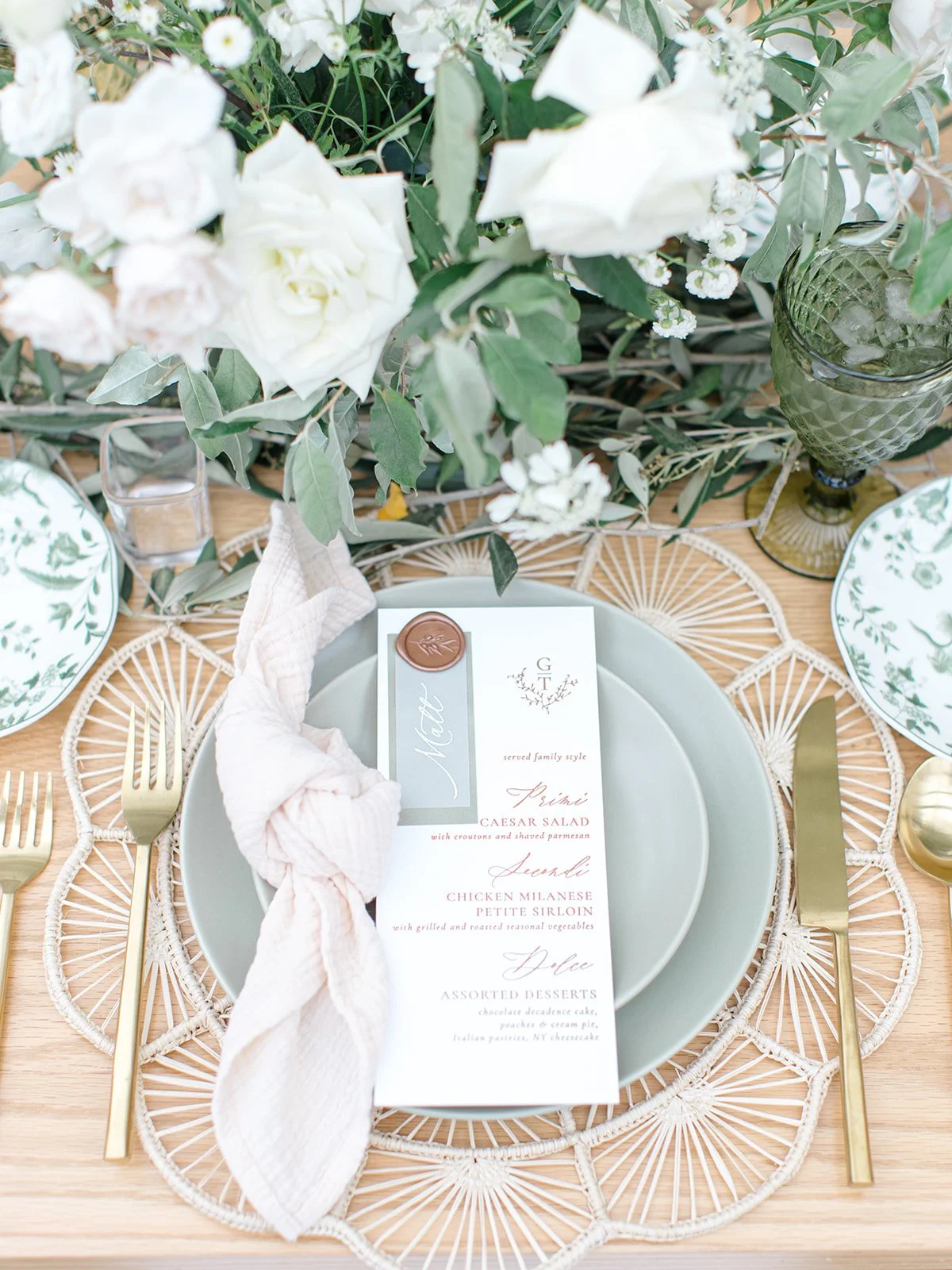 Elegant table setting with a light green plate, gold flatware, a white cloth napkin, a floral menu card, and a blurred floral centerpiece with white flowers and greenery.