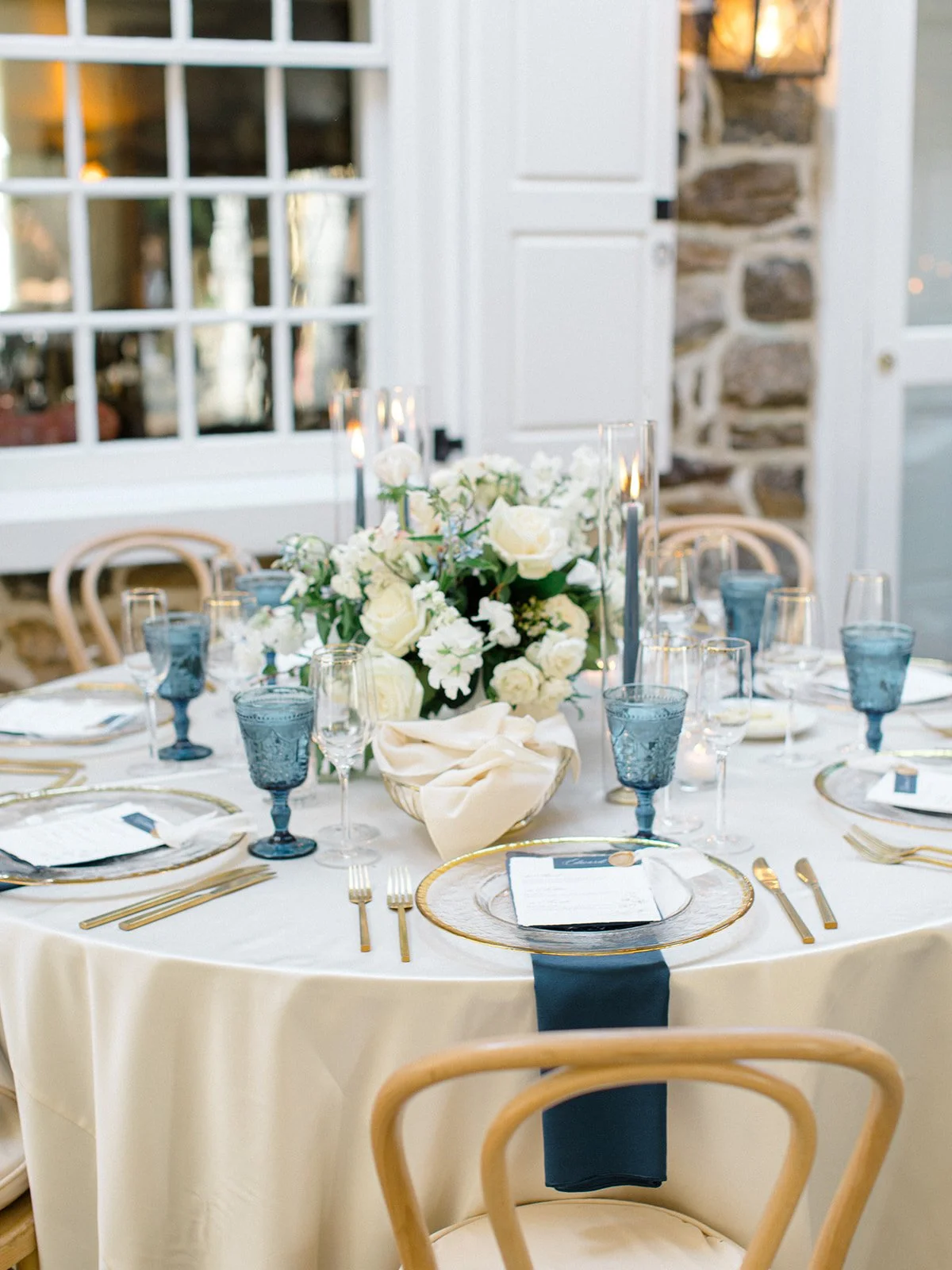 Round table set for a formal event with white tablecloth, gold cutlery, blue glasses, and a floral centerpiece of white roses and other white flowers, in a well-lit room with stone and white walls.