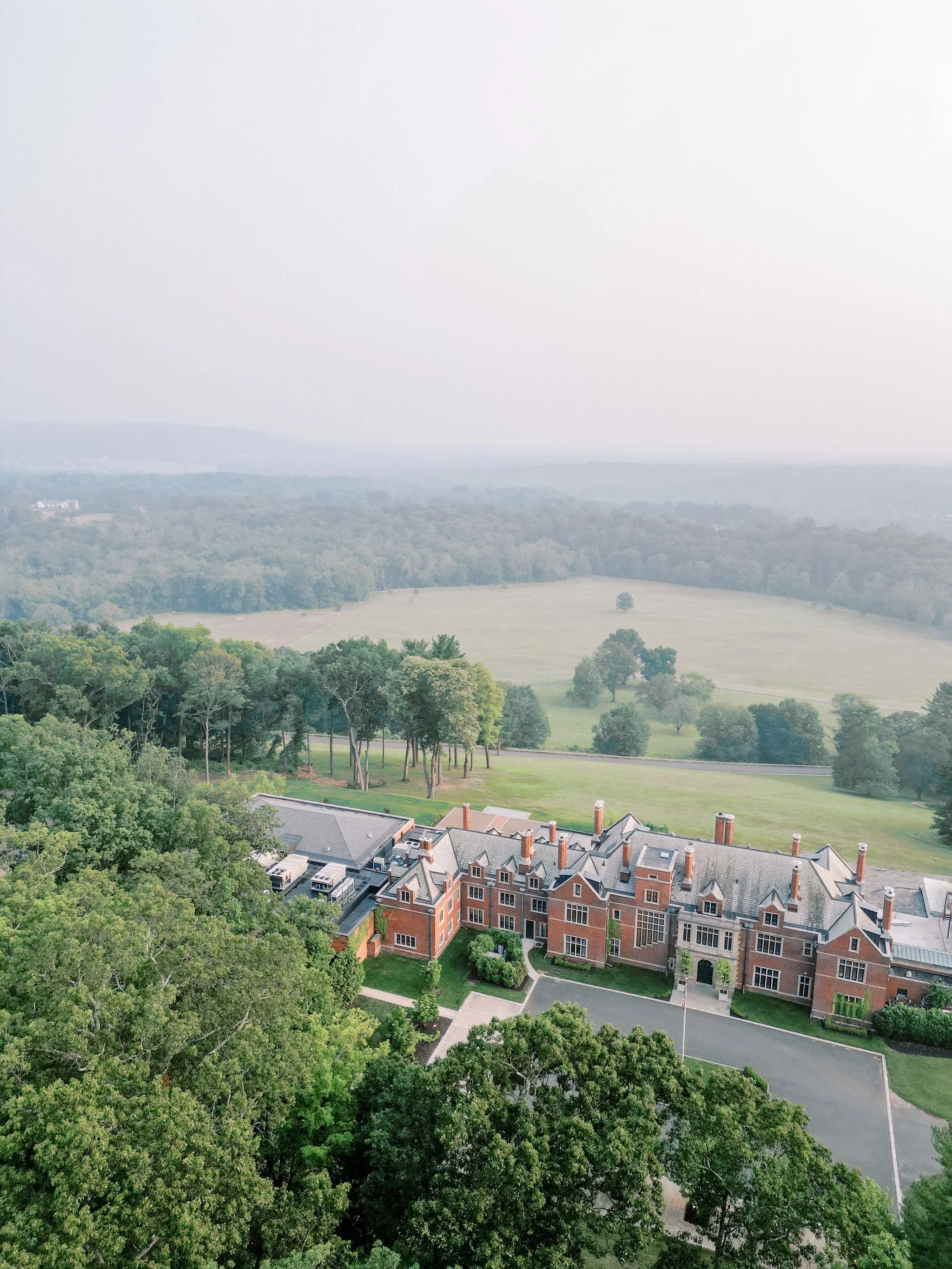 Aerial view of a large red brick mansion with a gray roof surrounded by green trees, with open fields and distant hills in the background.