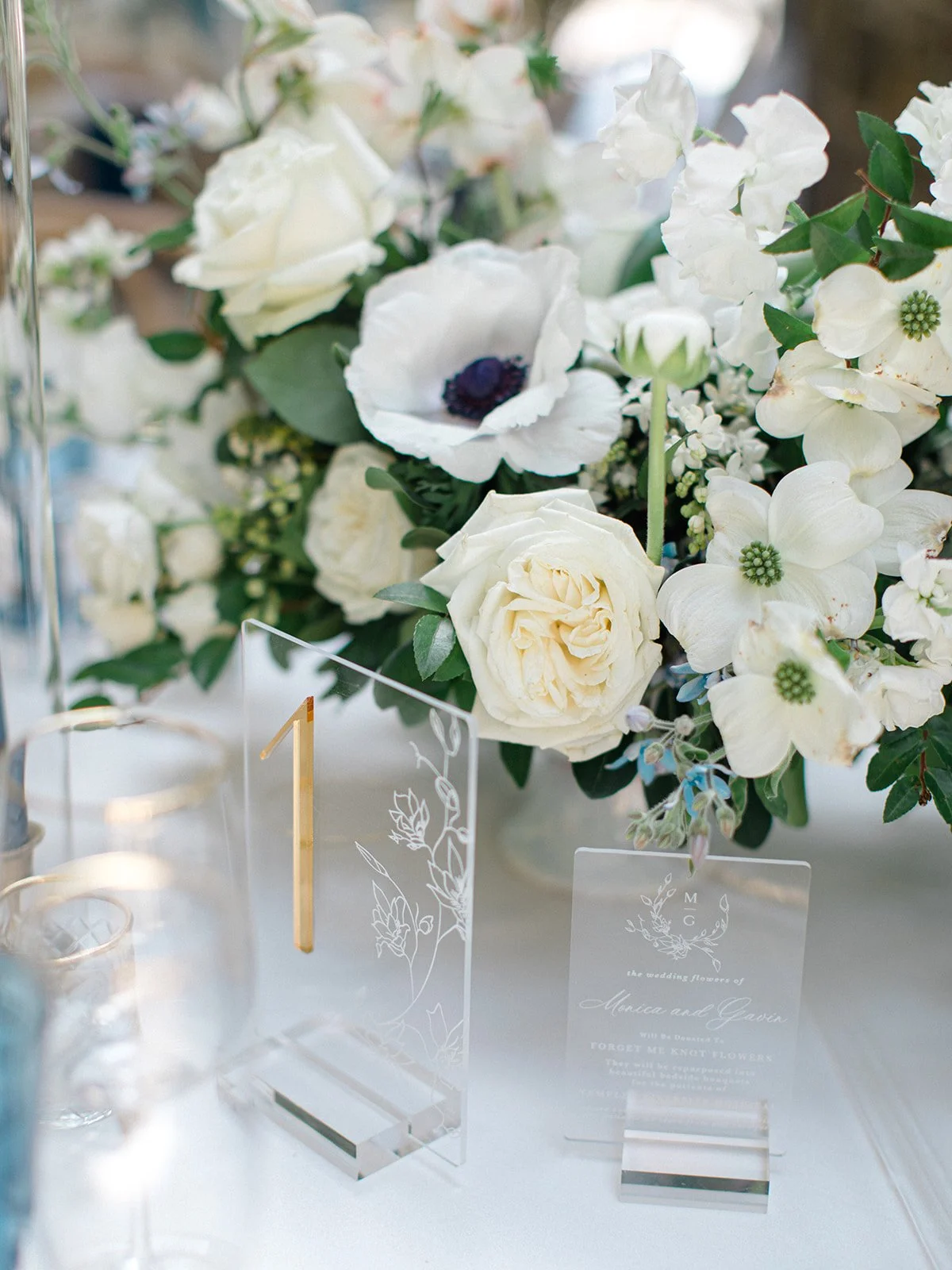Elegant wedding table centerpiece with white roses, anemones, hydrangeas, and greenery, accompanied by clear acrylic place cards and table number.