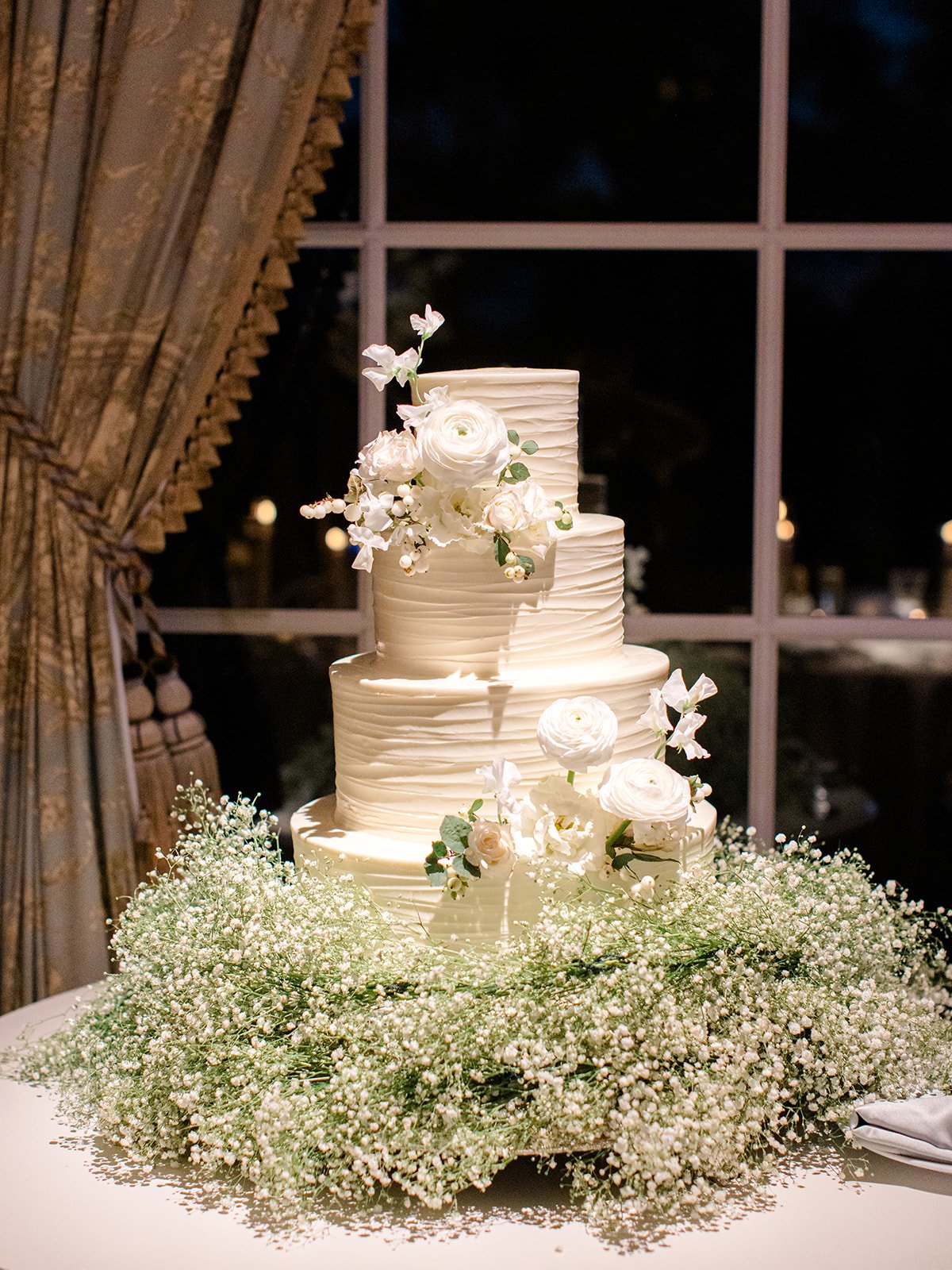 A multi-tiered white wedding cake decorated with white flowers, surrounded by baby's breath flowers, placed on a table in front of a window.