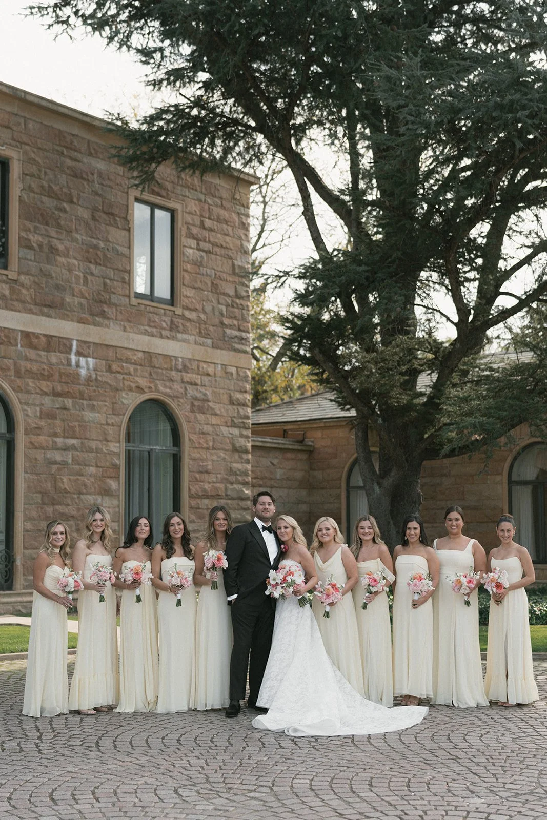 Wedding party with bride and groom surrounded by bridesmaids, standing outdoors on stone-paved area. The bride wears a white wedding gown, and the groom is in a black tuxedo. Bridesmaids are in cream-colored dresses, holding bouquets of pink and whit