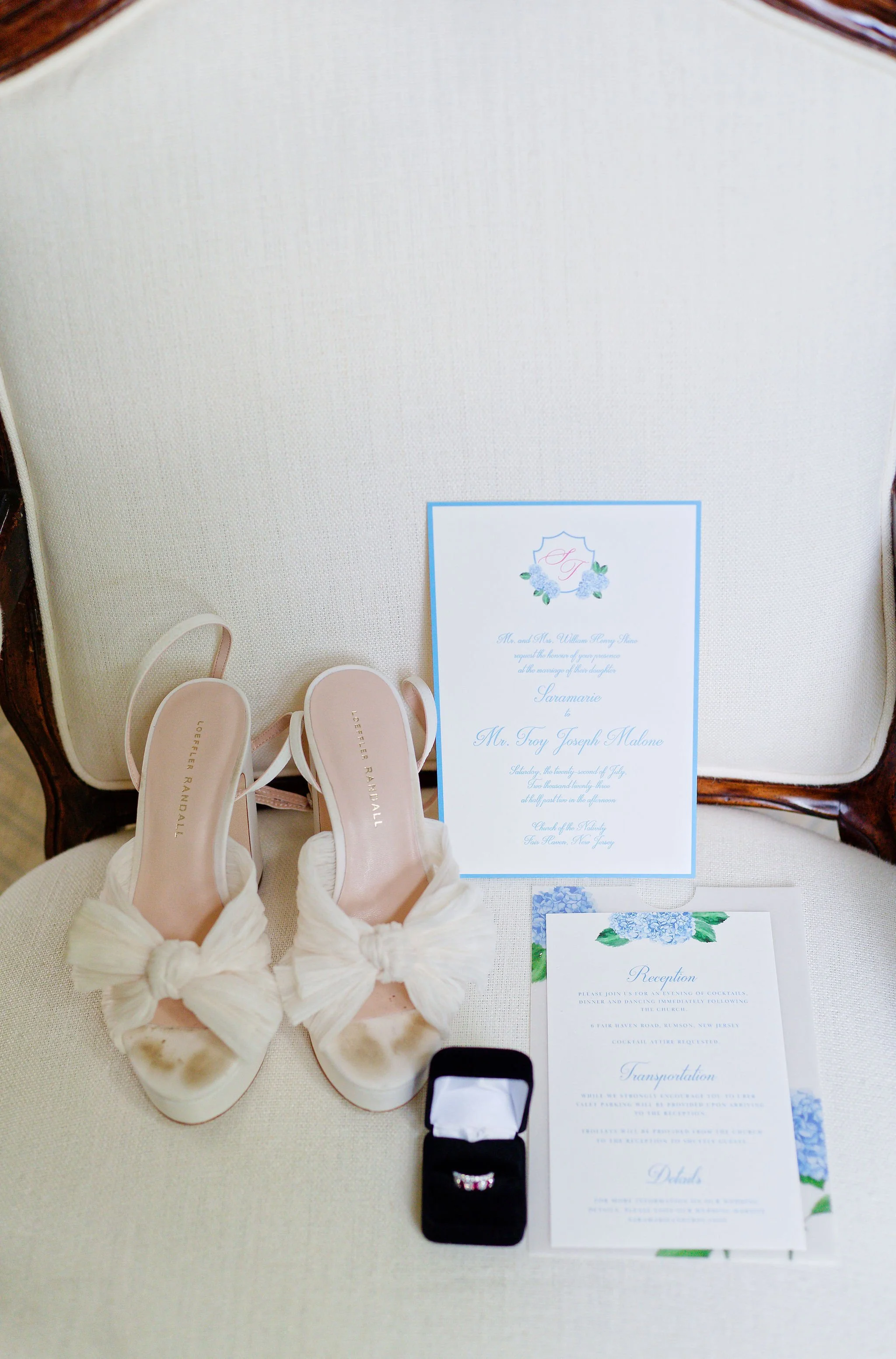 Wedding shoes, invitation cards, a wedding ring in a jewelry box, and a reception program on an elegant chair