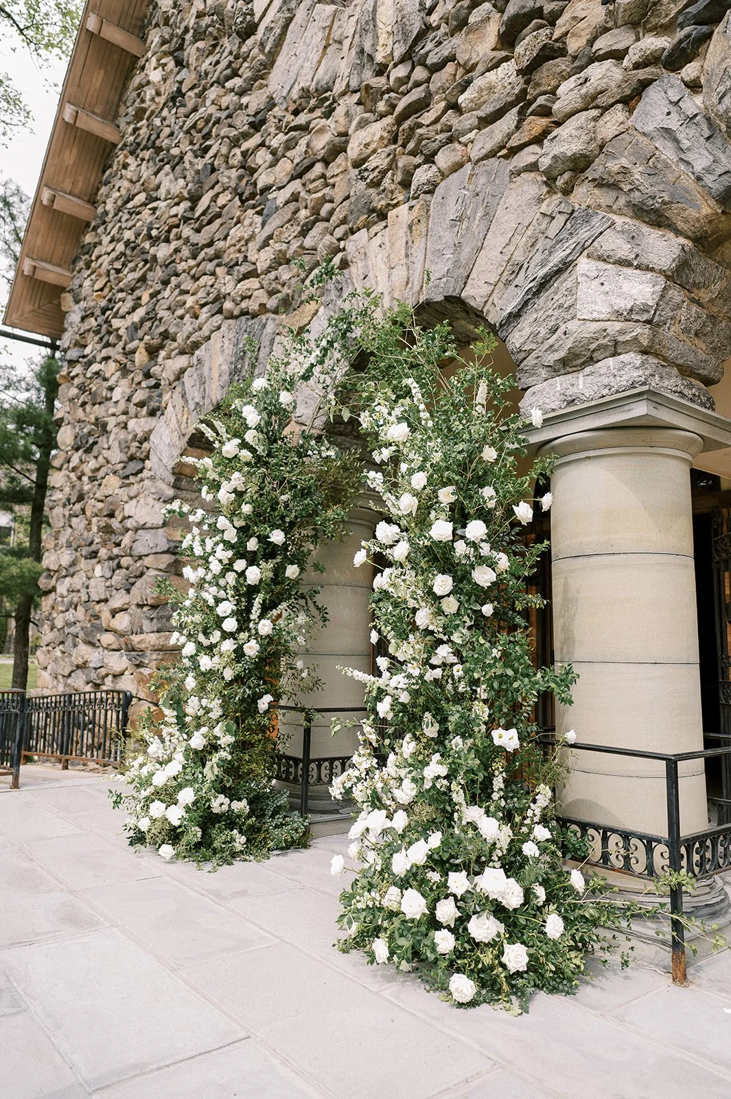Flower arch with white roses and greenery outside a stone building with columns.