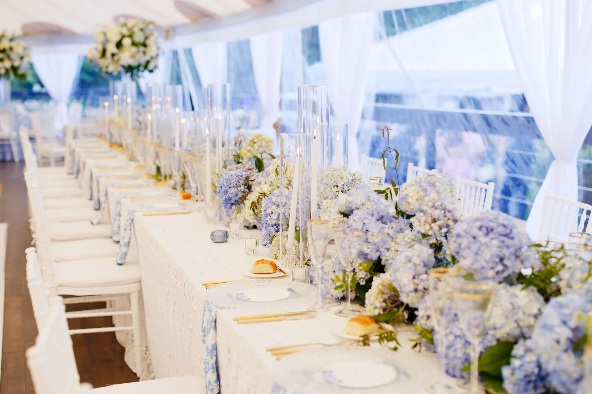 Elegant wedding reception table decorated with white and blue hydrangea flowers, tall glass candle holders, gold-rimmed glassware, white plates, and gold cutlery, set inside a bright tent with white curtains.