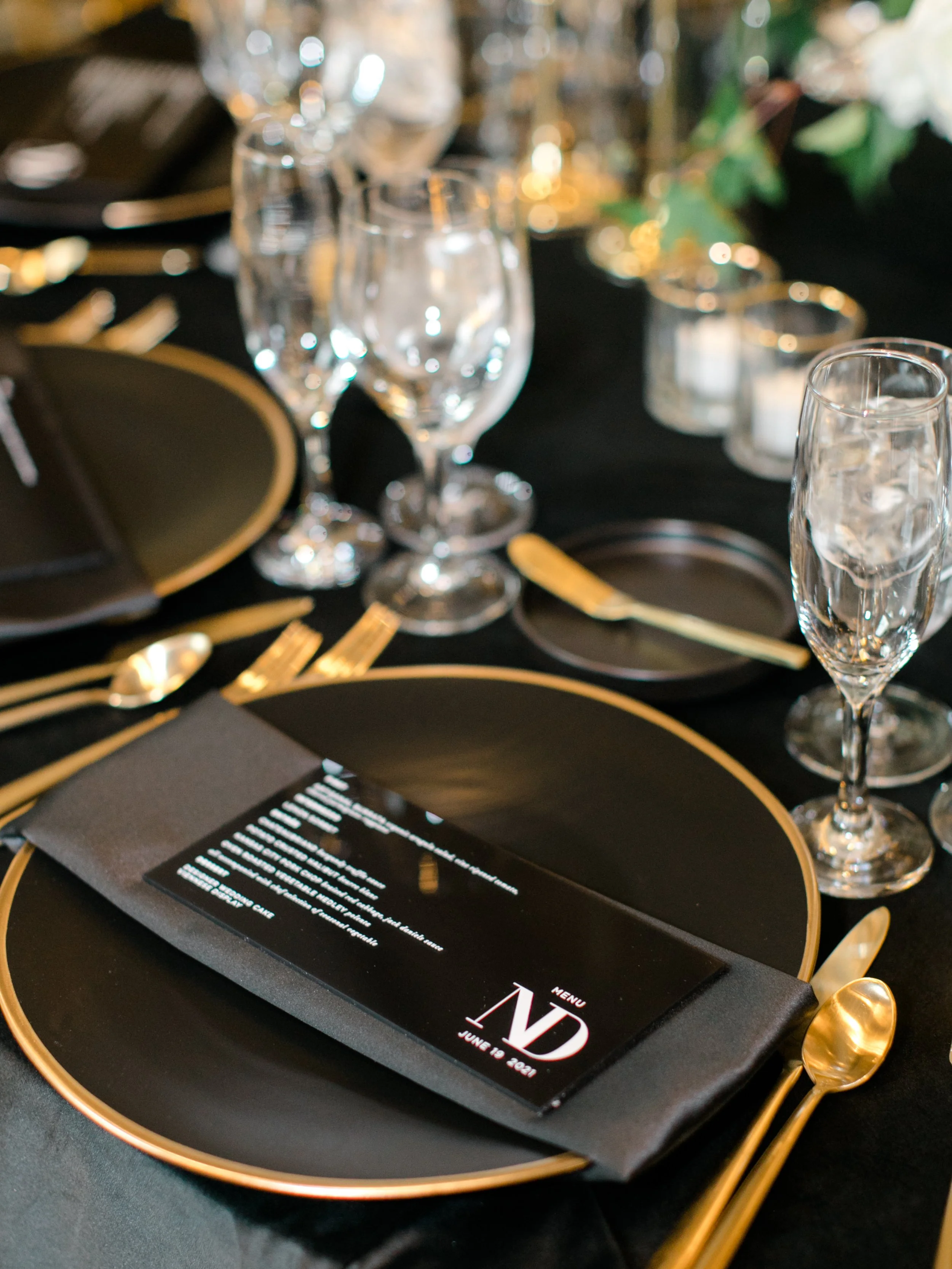 Elegant dinner table setting with black and gold plates, gold utensils, and glassware, including wine glasses. A black menu card with white and red text is placed on a black napkin.