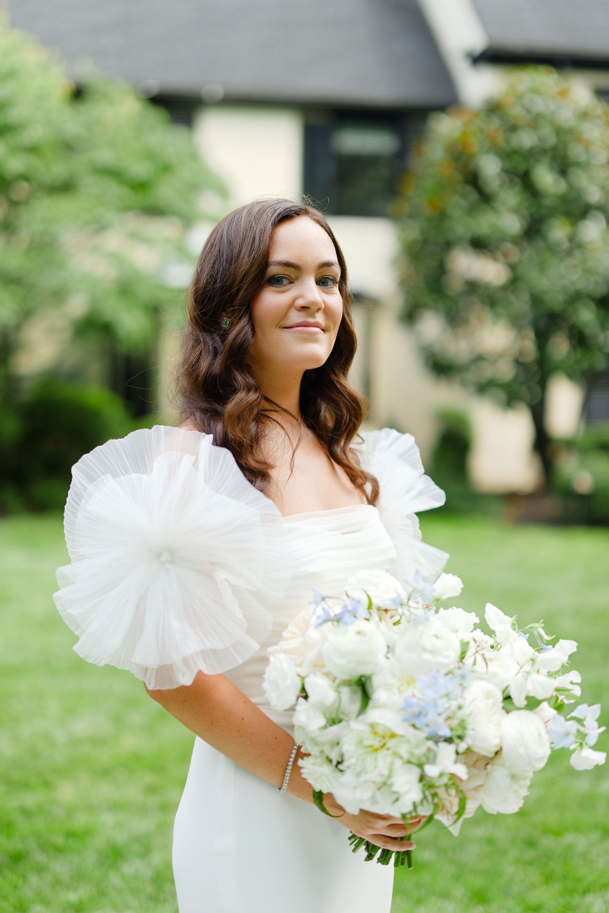 A woman in a white dress holding a bouquet of white flowers outdoors with trees and a house in the background.