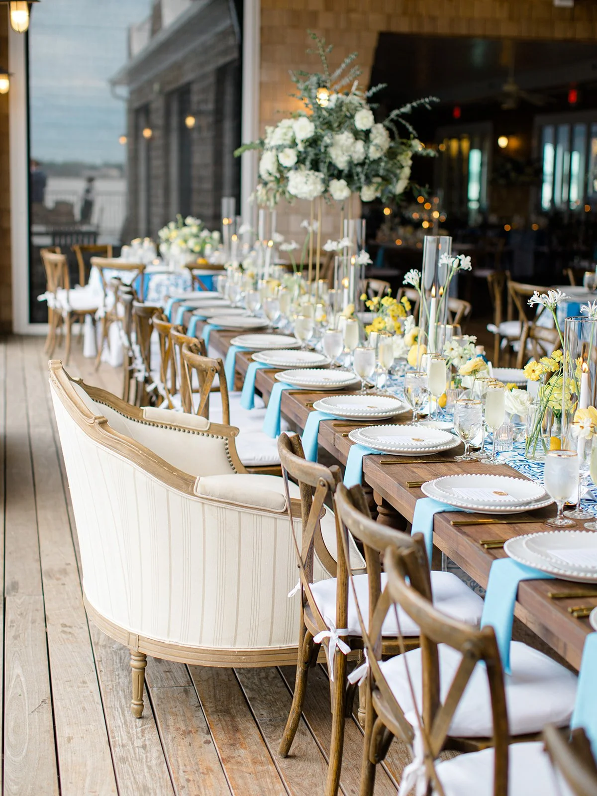 Elegant banquet table decorated with white and yellow flowers, blue table runners, and glassware, set for a formal event on a wooden porch.