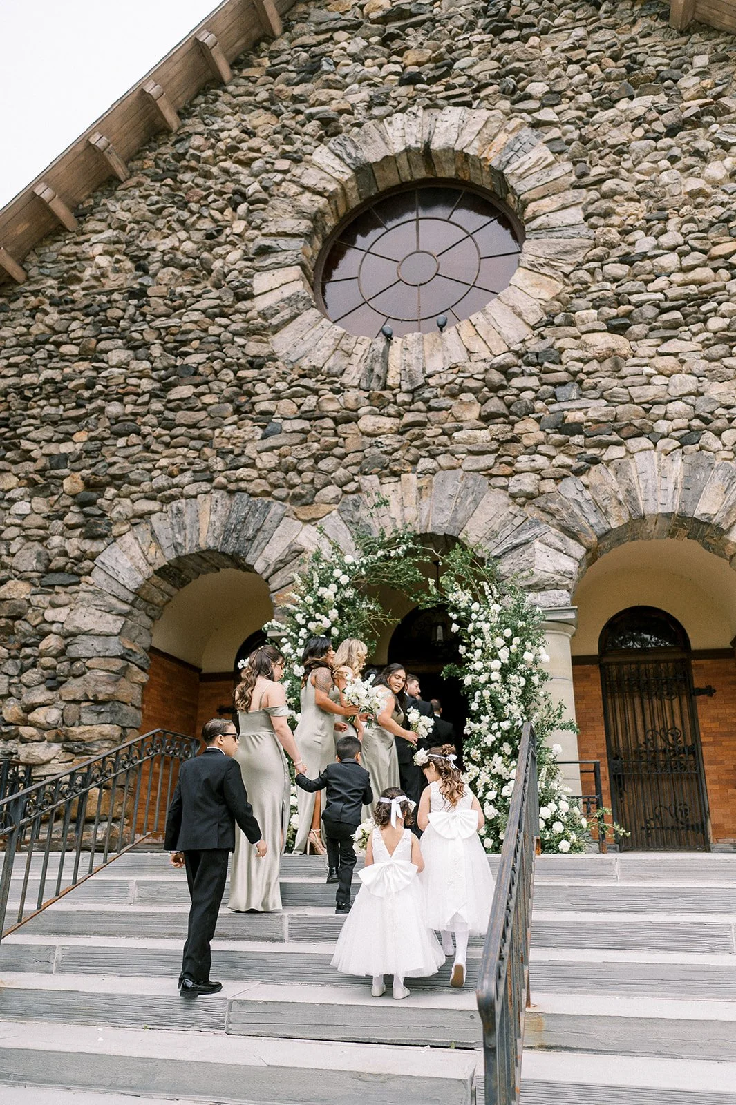 A wedding party, including children in white dresses, ascending the steps of a stone church decorated with white flowers and greenery, preparing for the ceremony.