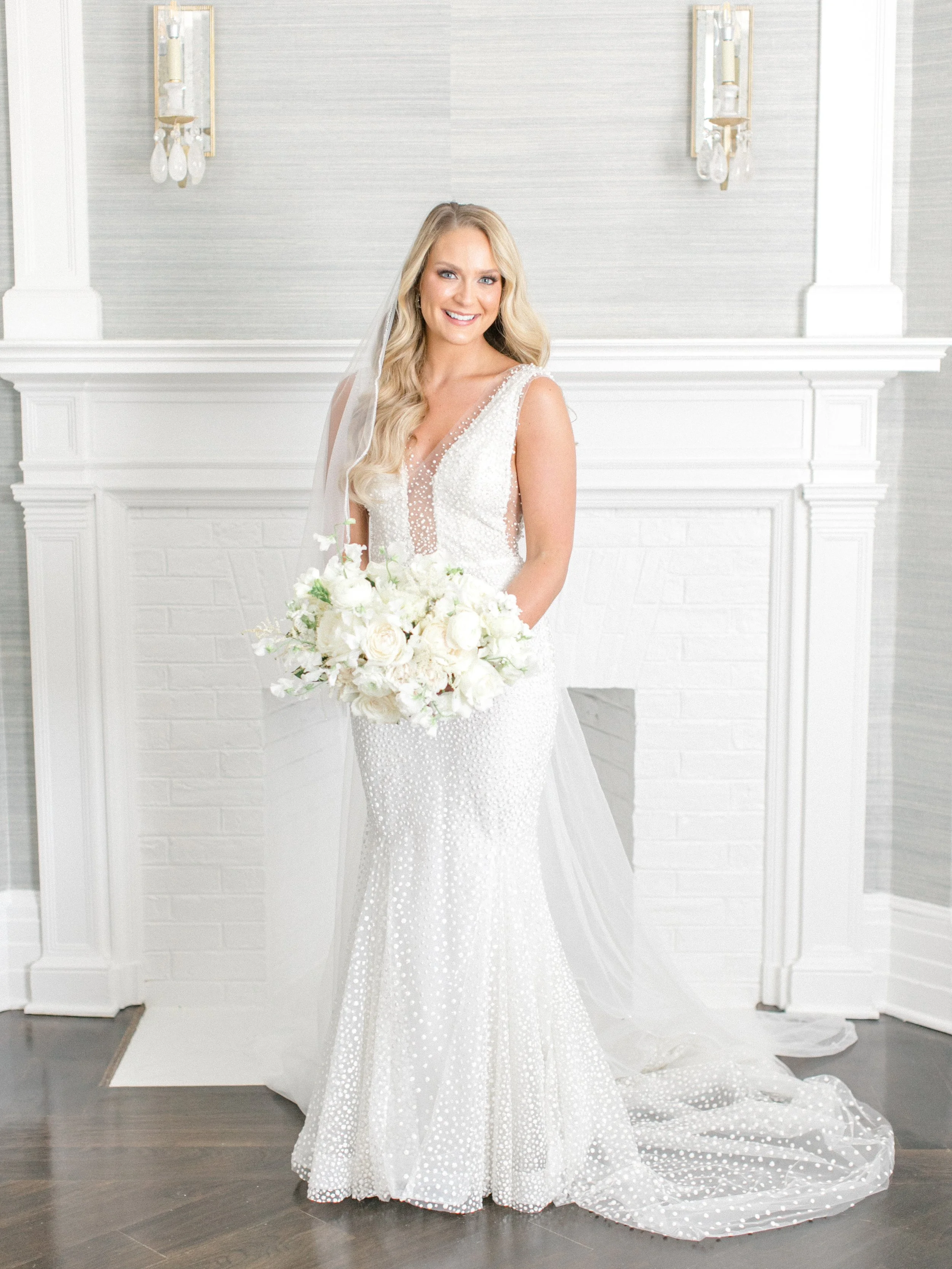 A bride in a white wedding gown with bead embellishments, standing in front of a white fireplace with a bouquet of white flowers, smiling.