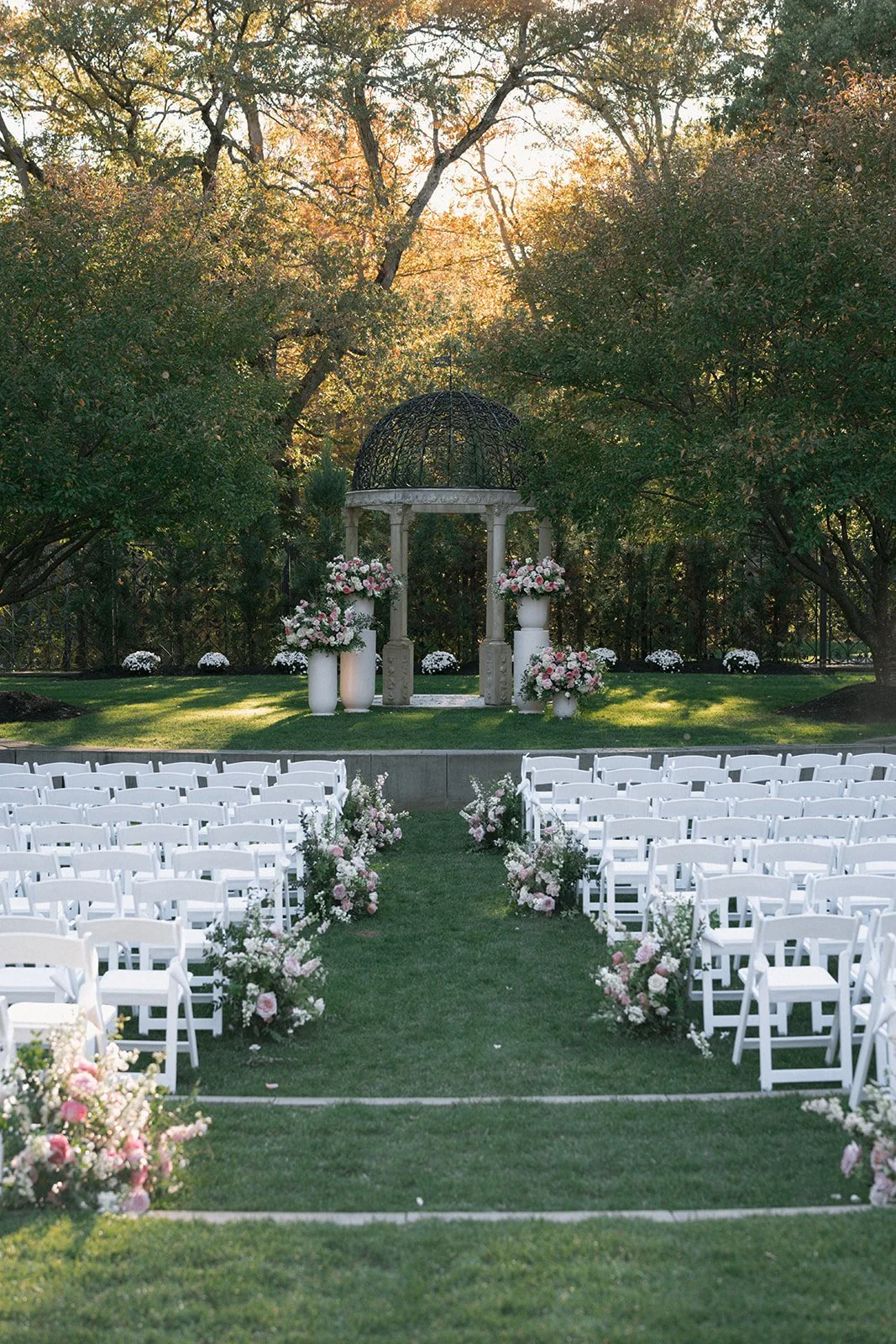 Outdoor wedding ceremony setup with white chairs arranged on either side of a green grass aisle, decorated with pink and white floral arrangements. At the end of the aisle is an altar with large floral bouquets and a small arched structure with a bla