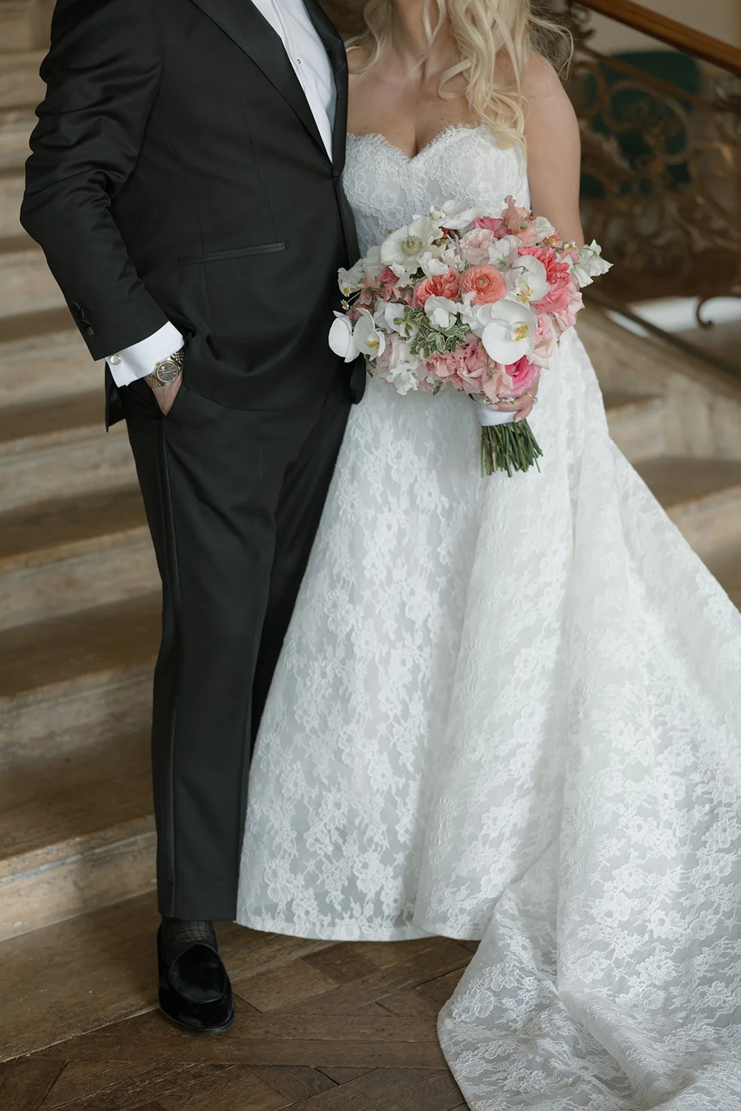 Bride and groom standing together on a staircase, with the bride holding a bouquet of pink and white flowers, wearing a white lace gown and the groom in a black suit with a white shirt.