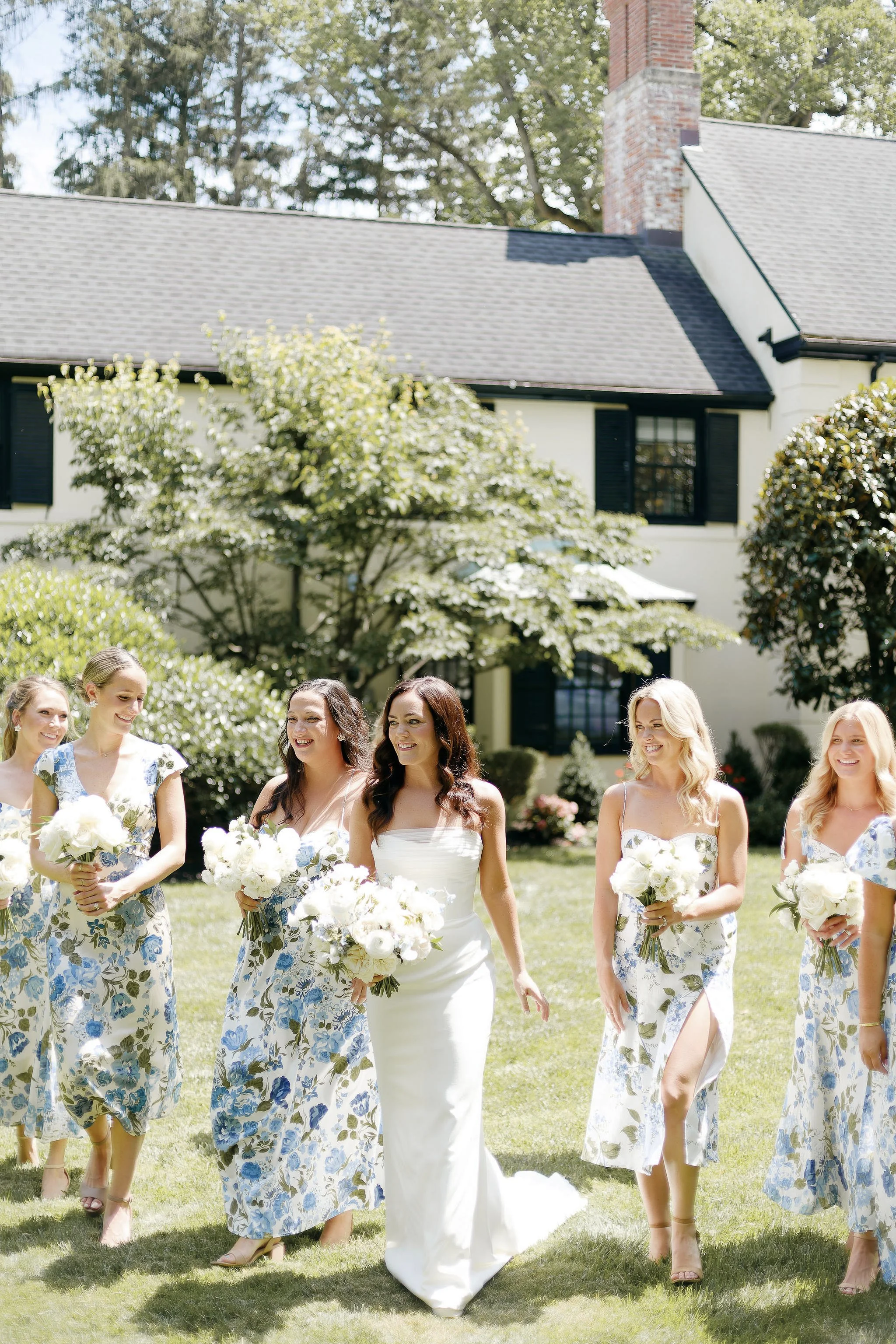 A bride in a white wedding dress walking outdoors with five bridesmaids in floral dresses, holding bouquets, on a sunny day in front of a house with a gray roof, white walls, and green trees.