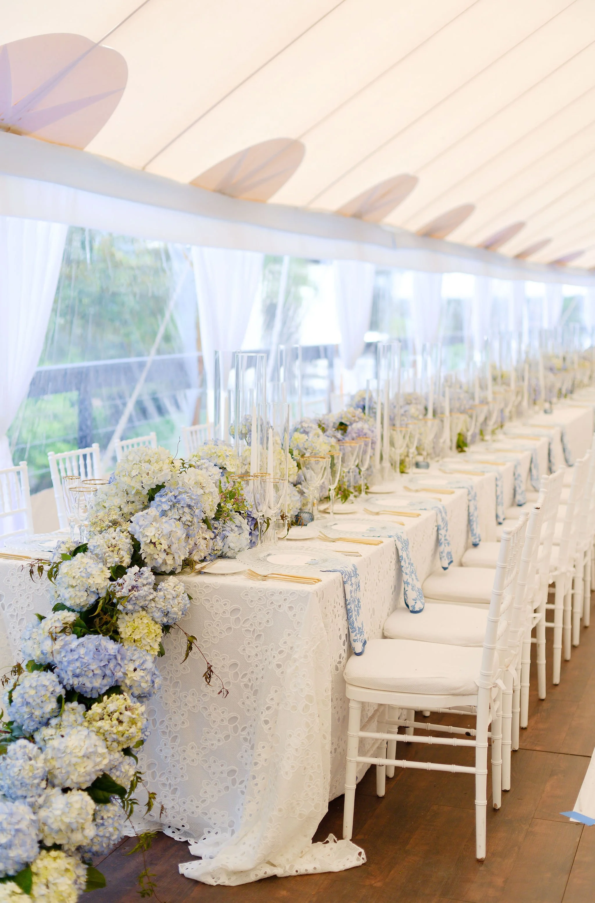 Elegant white banquet table decorated with pastel blue and white floral arrangements, surrounded by white chairs, set up for a wedding reception or special event in a tented venue with sheer curtains.