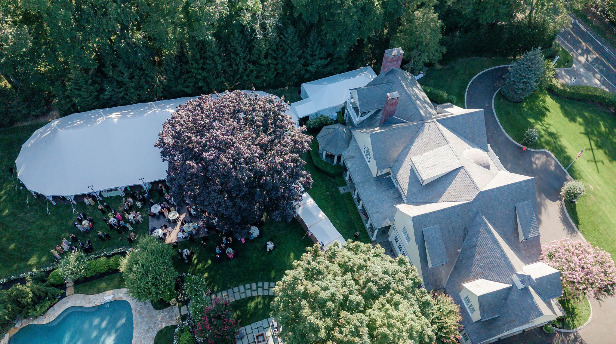 Aerial view of a large house with a garden and multiple trees, a curved driveway, and an outdoor event with tents and people gathered around tables.