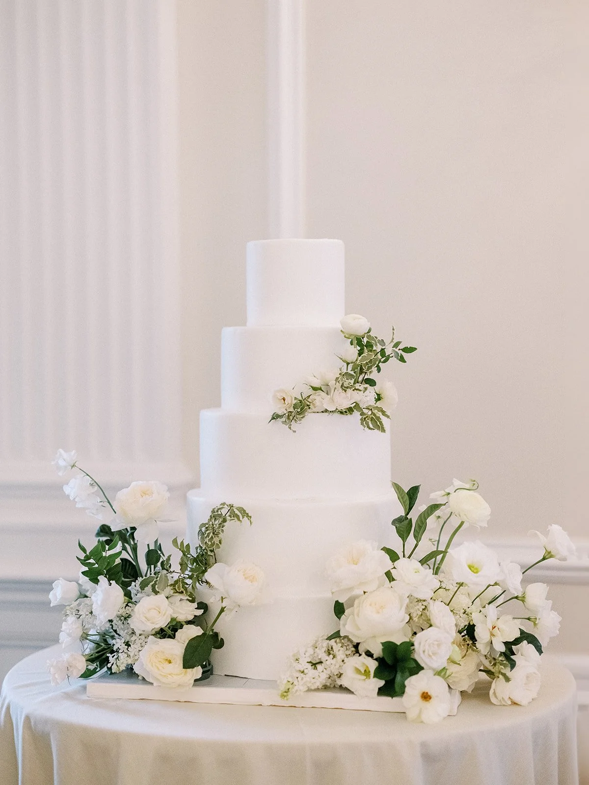 White wedding cake with four tiers decorated with white flowers and green foliage, placed on a round table with a white tablecloth.