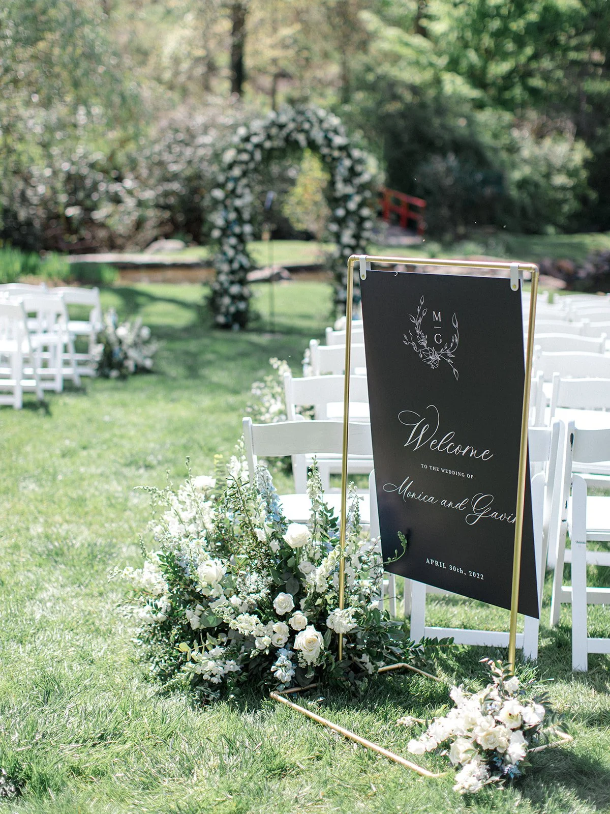 Outdoor wedding setup with white chairs, a floral arch in the background, and a welcome sign for Monica and Gavin's wedding on April 30th, 2022, decorated with white flowers and greenery.
