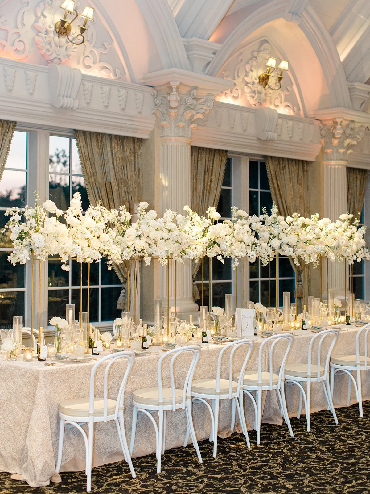 Elegant banquet table decorated with white flowers, candles, and glassware in a luxurious room with ornate moldings, large windows, and gold accents.