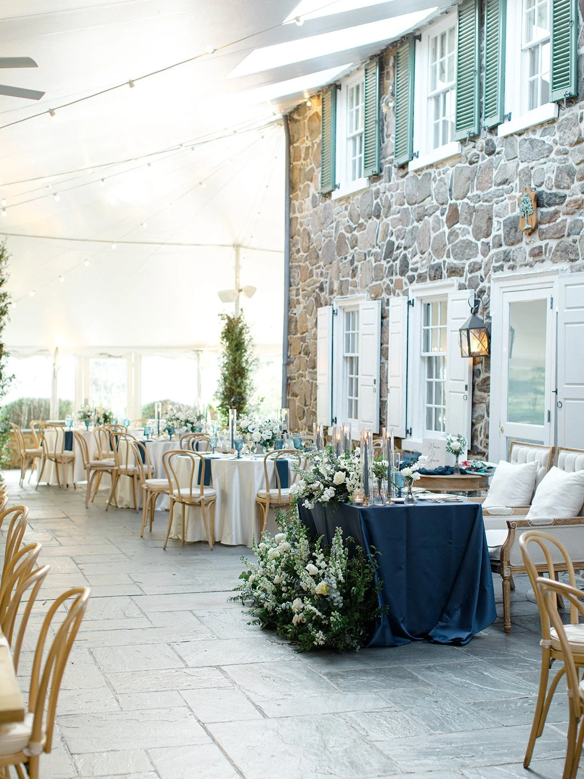 Elegant outdoor wedding reception with round tables covered in white tablecloths, surrounded by wooden chairs, and decorated with floral centerpieces and candles. A stone building with white windows and shutters is visible in the background, and stri