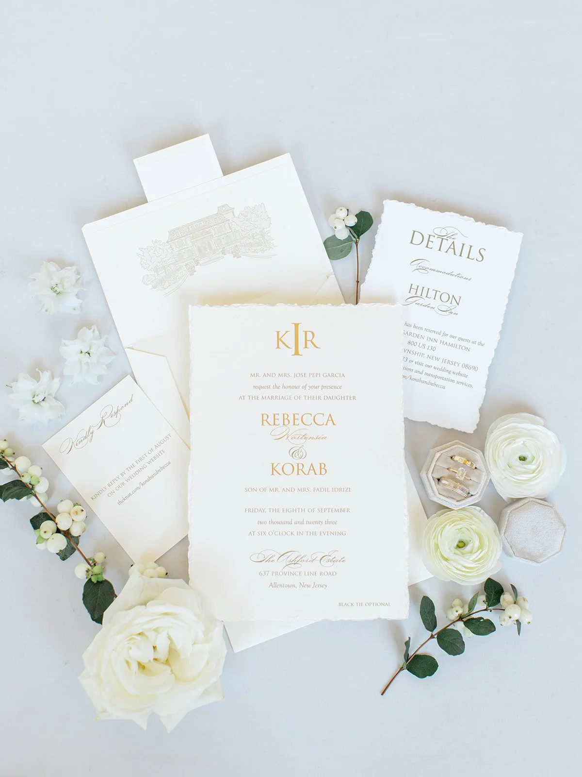 Wedding invitation with gold details, surrounded by white flowers, rings, and green leaves on a light-colored surface.