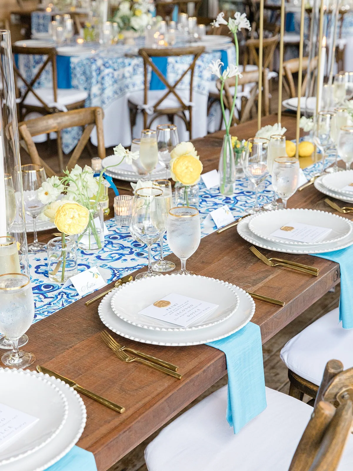Elegant dining table set for a formal event with white plates with beaded edges, gold flatware, blue cloth napkins, water glasses with gold rims, small vases with yellow and white flowers, and a blue and white patterned runner.