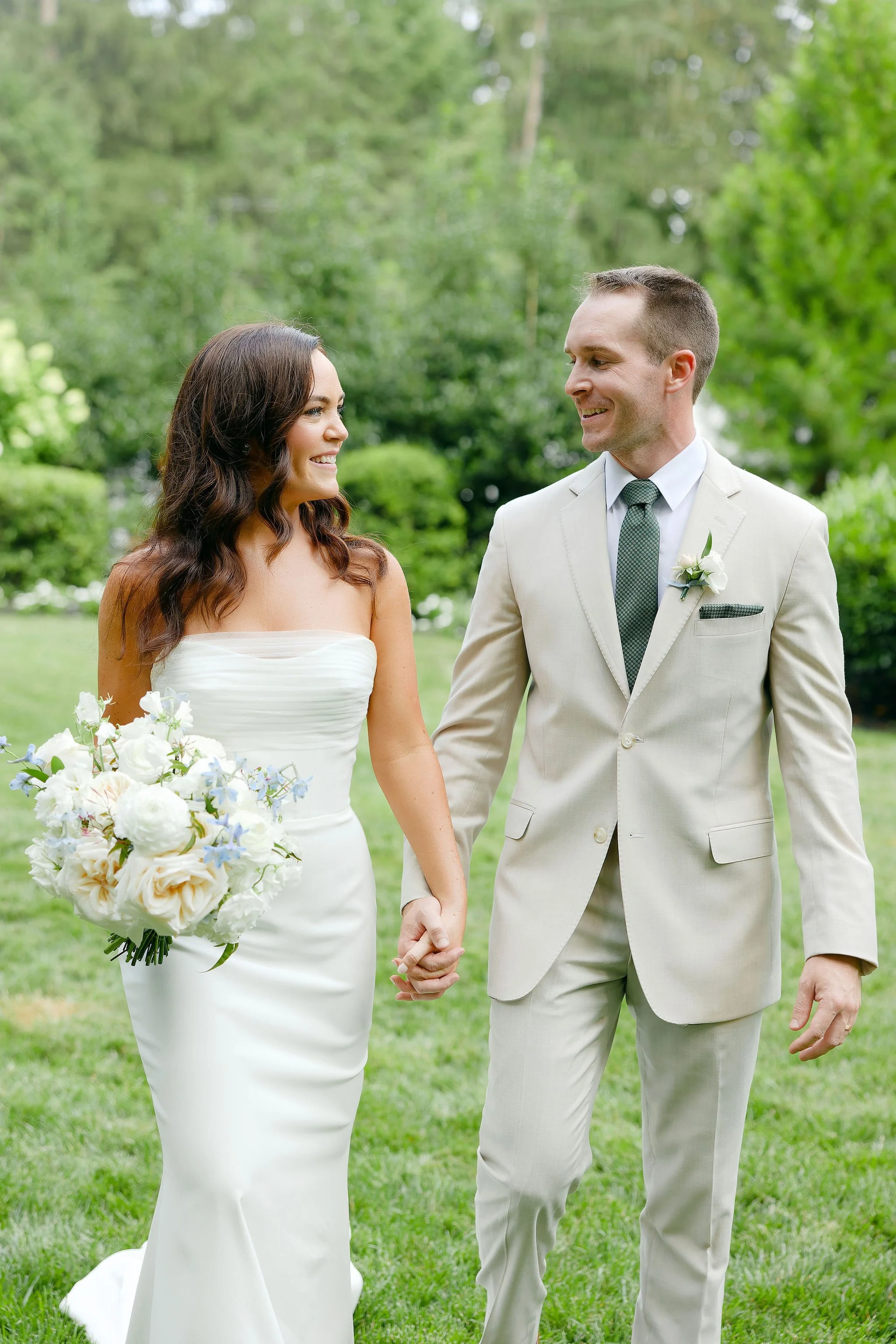 A bride and groom holding hands outdoors, smiling at each other, with greenery in the background.